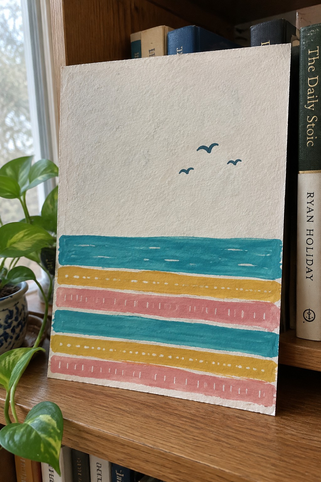 Colorful striped watercolor painting with birds on bookshelf beside plant and books.