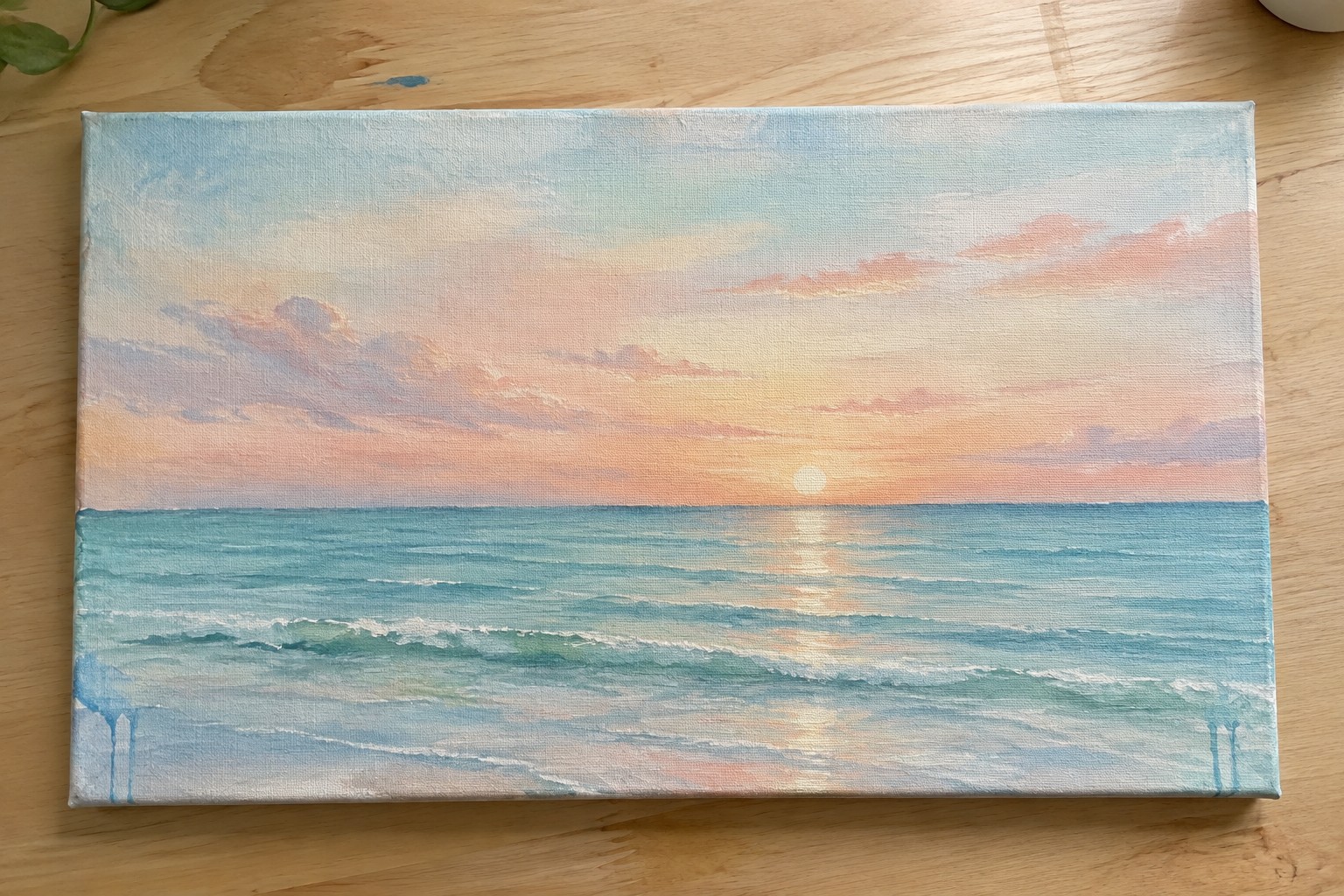 Colorful sunset over ocean waves painted on canvas atop wooden table with plant.