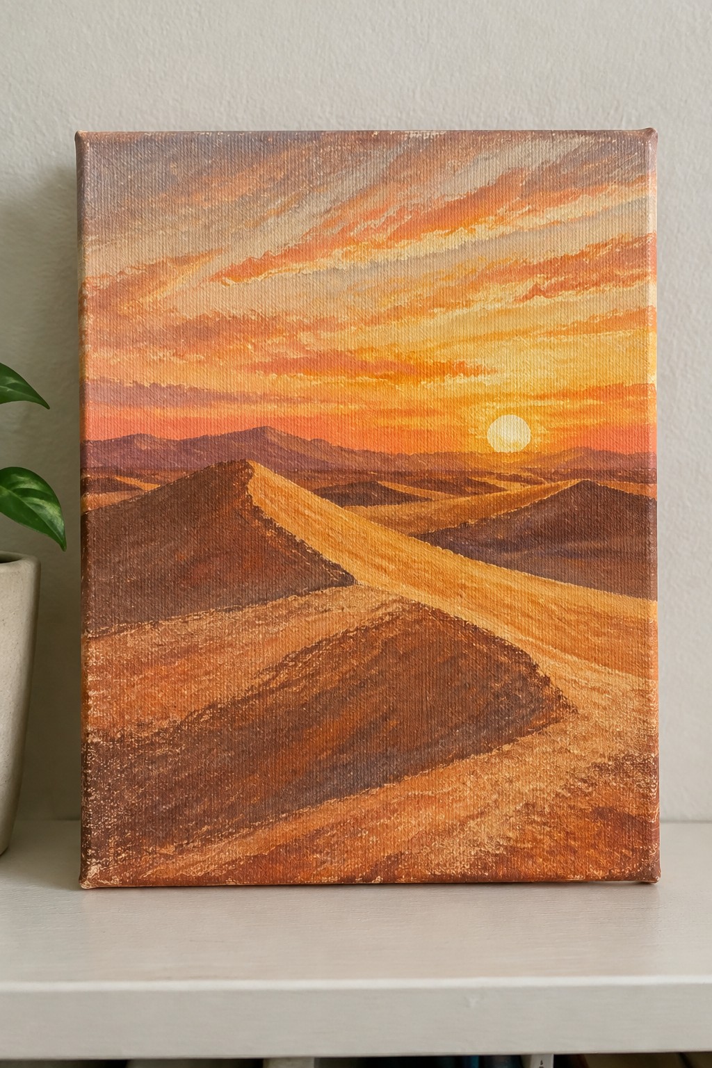 Painting of desert dunes at sunset with orange sky, sun, and distant mountains