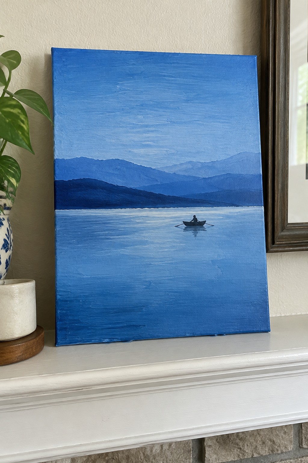 Blue mountain lake painting with rowing boat on white mantel beside potted plant.