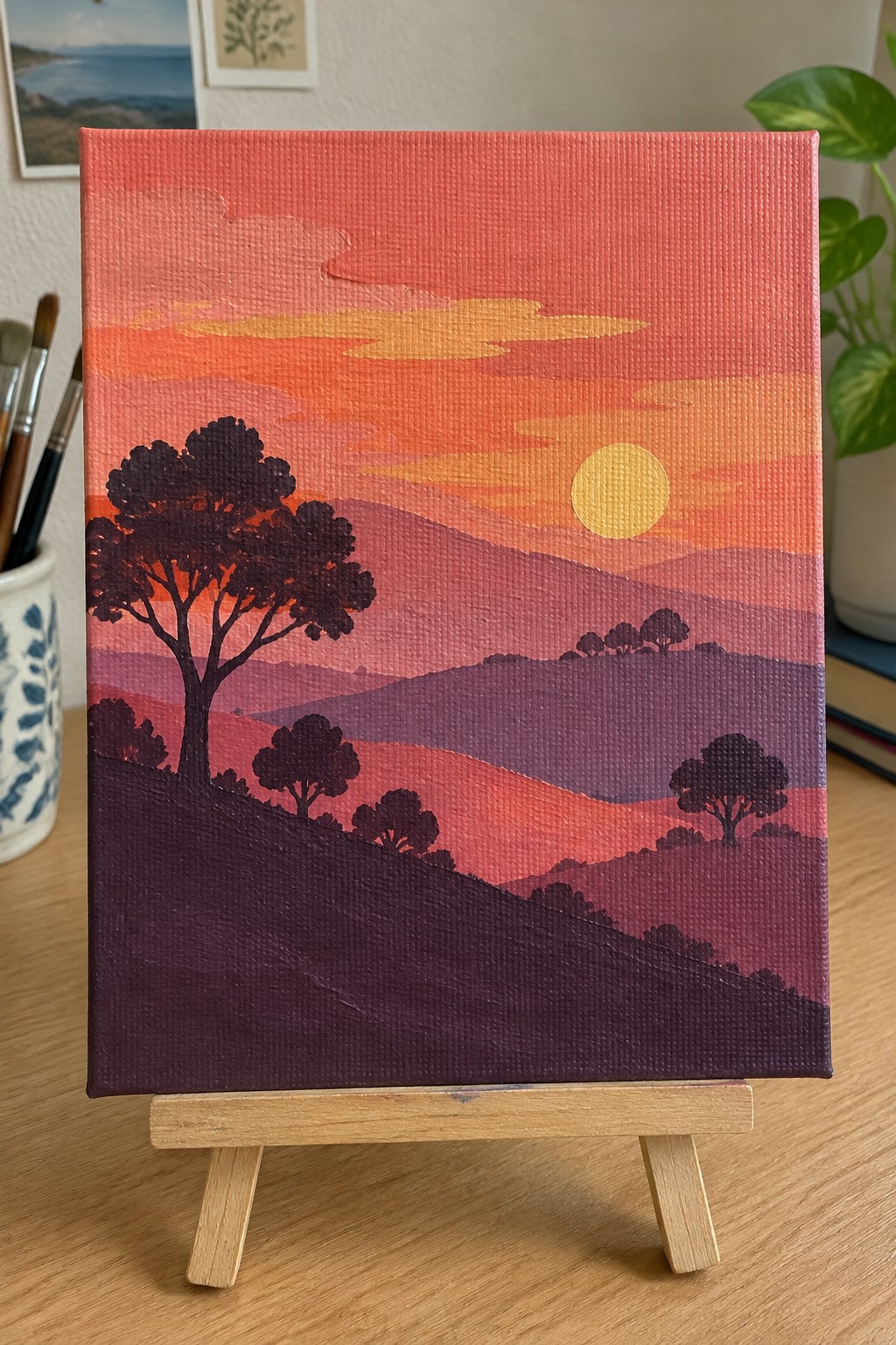 Vibrant sunset landscape painting with layered hills, silhouetted trees, and yellow sun on easel.