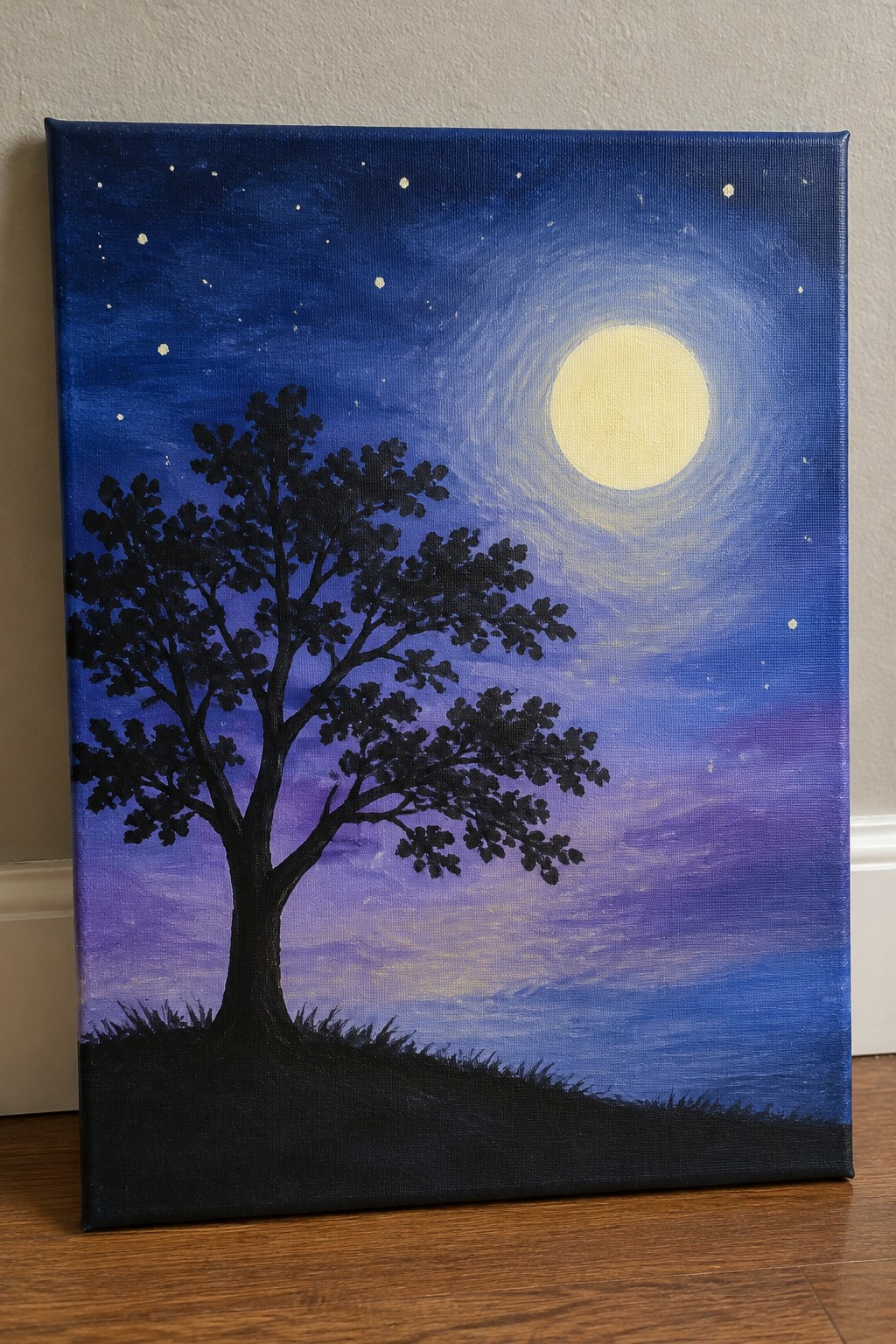 A canvas painting showing a black tree silhouette in front of a large moon and stars on a blue-purple gradient sky with dark ground.