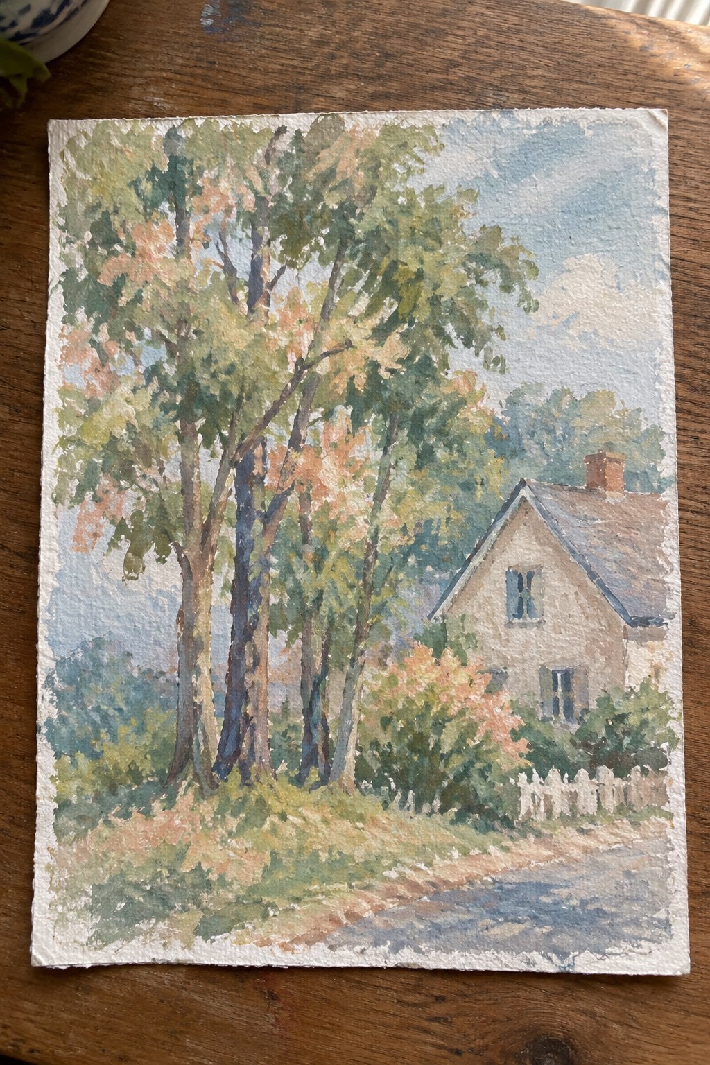 A watercolor landscape shows a light house with a dark roof on the right, tall trees with green and orange leaves on the left, a dirt path in front, and a white picket fence near the house.