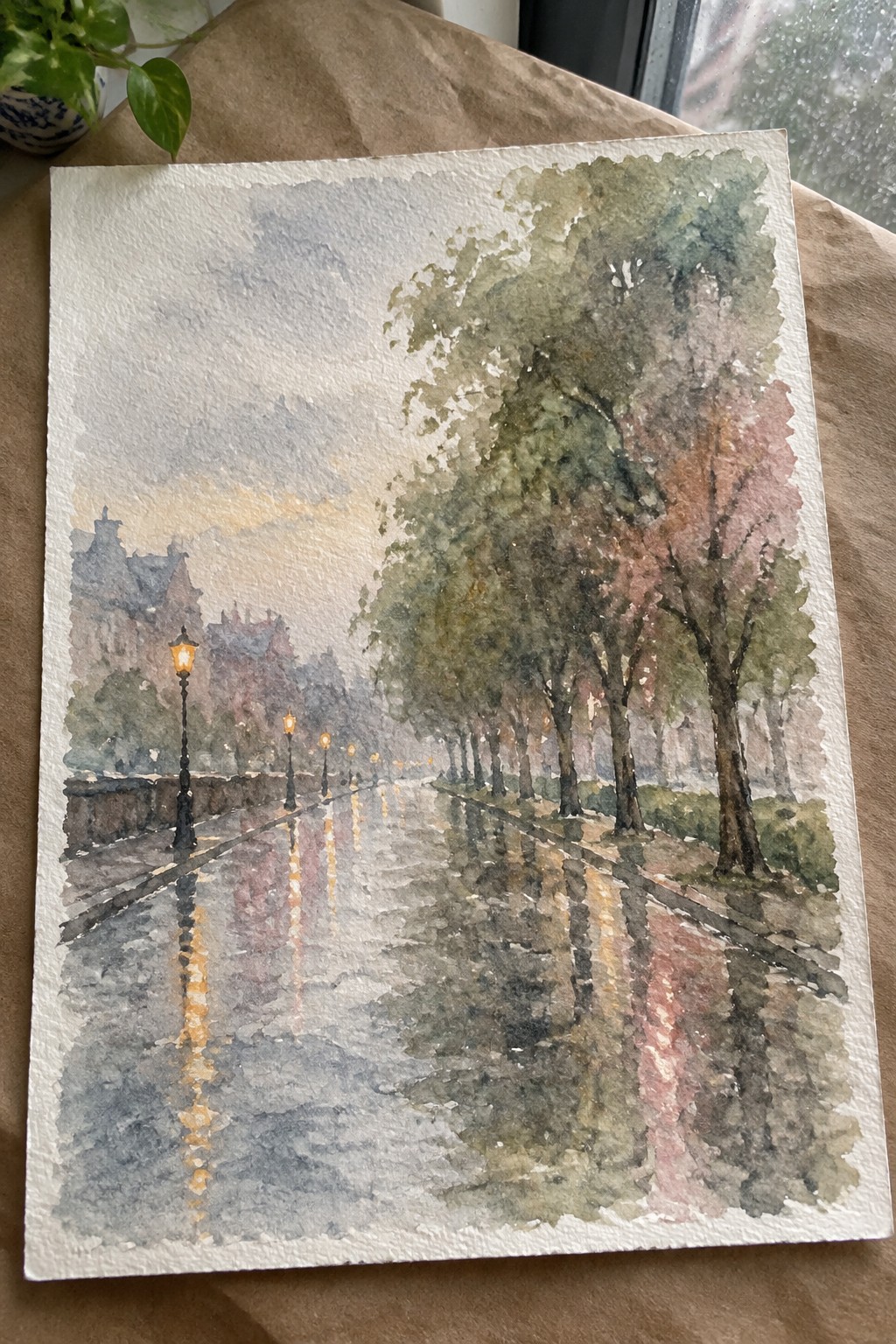 Watercolor of rainy tree-lined street with glowing lampposts reflecting on wet pavement.