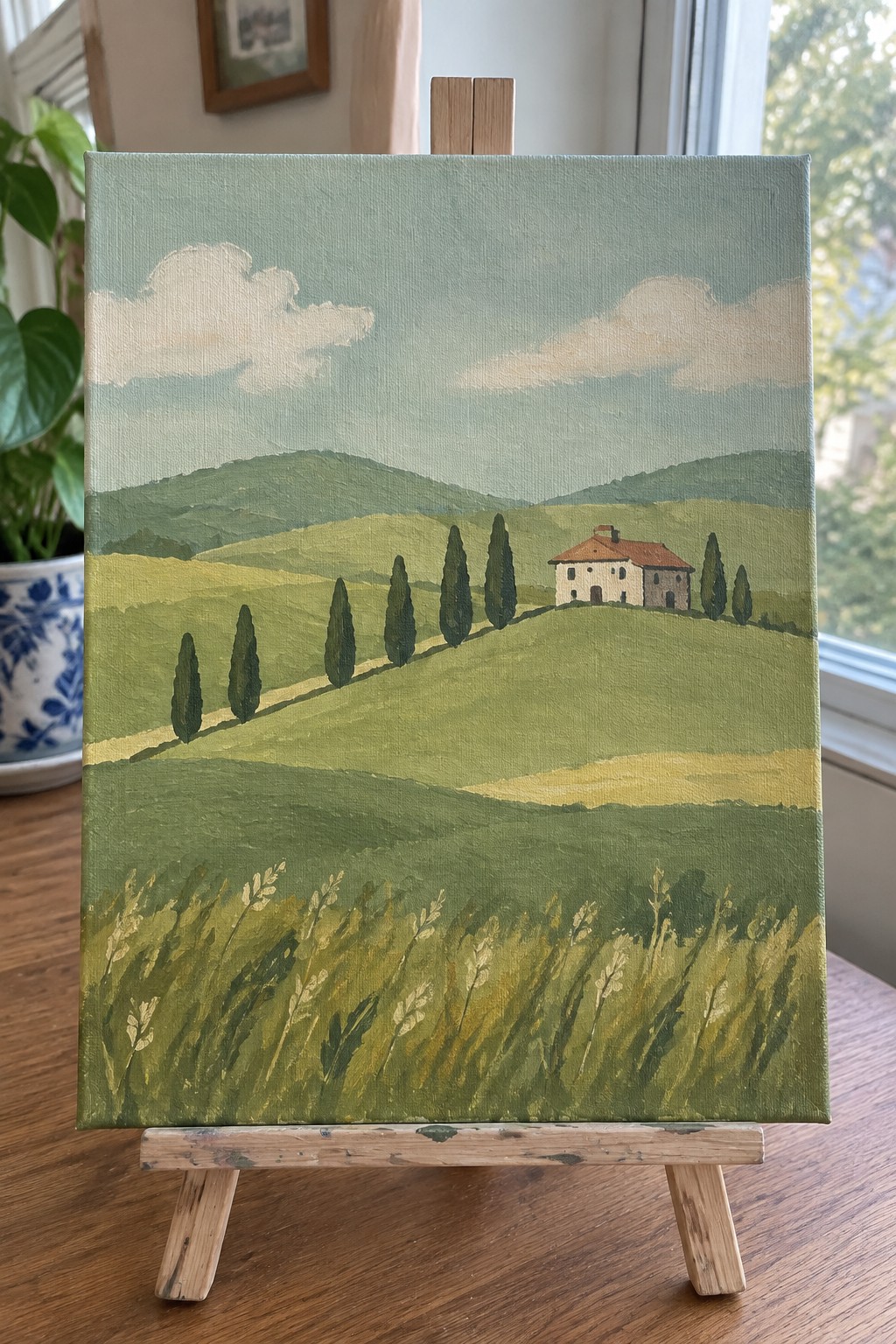 Landscape painting of rolling green hills, cypress trees, and red-roofed house on wooden easel