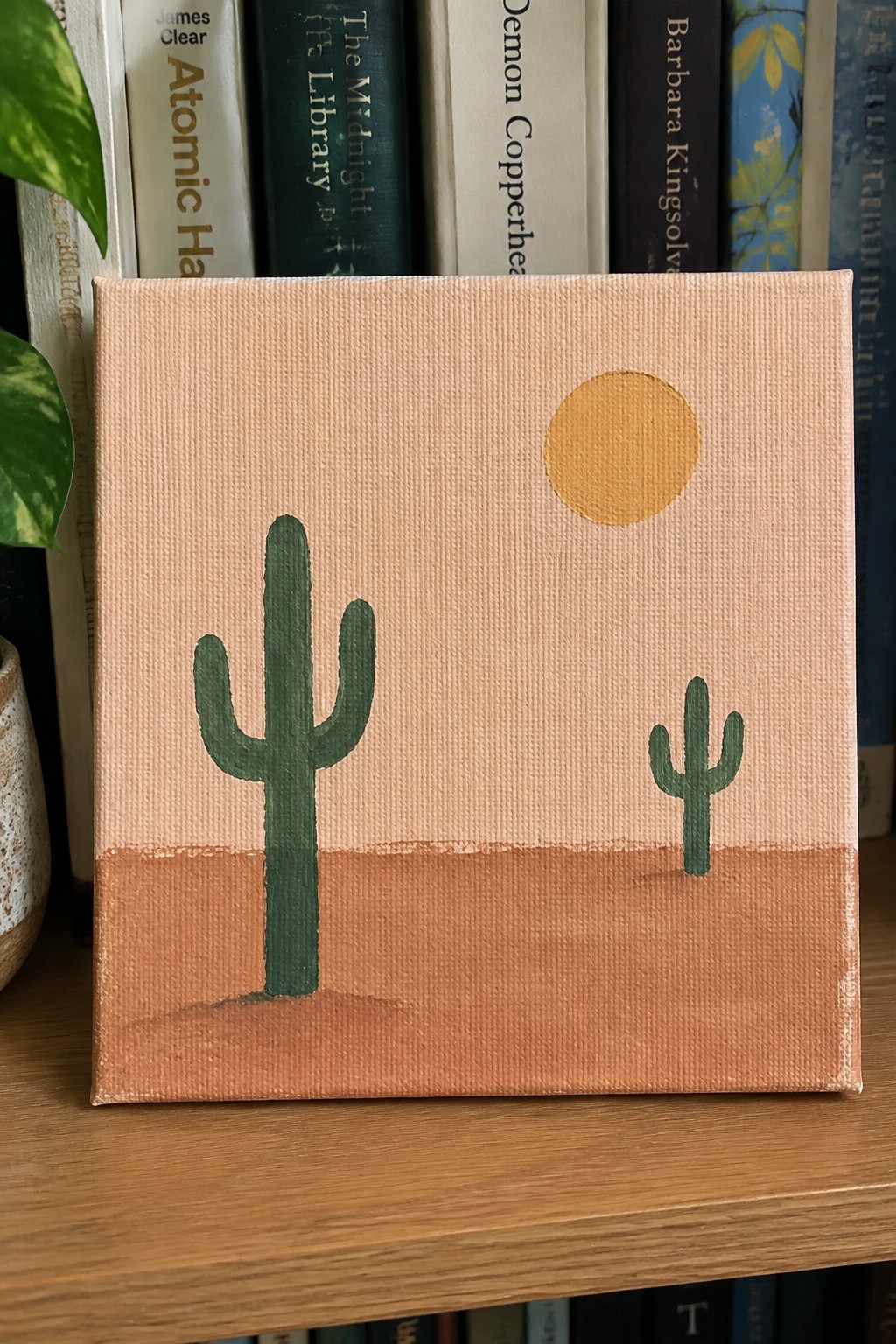 Small canvas desert painting with two green cacti, yellow sun, peach sky.