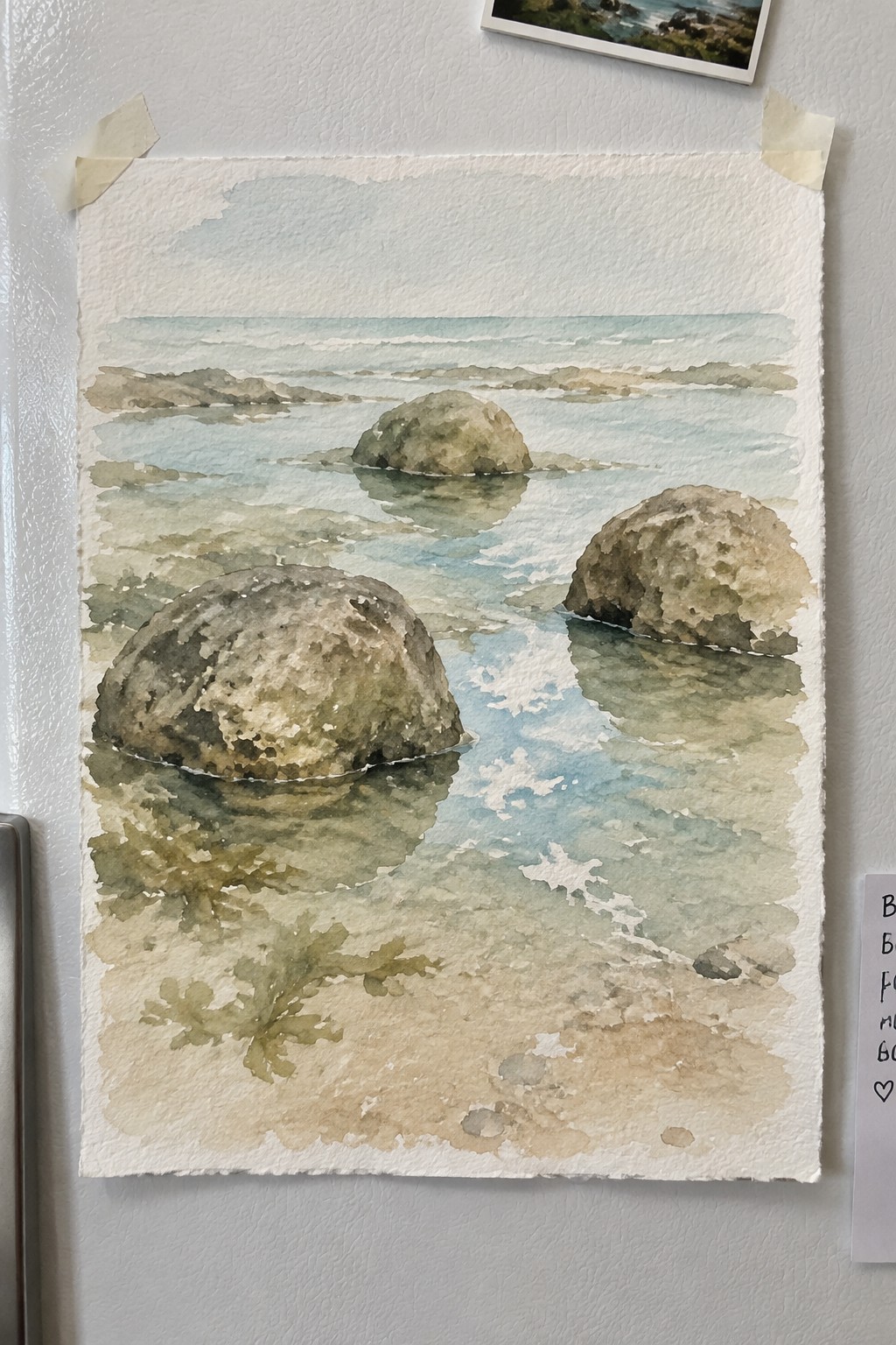 Watercolor seascape of boulders in shallow water taped to white fridge.