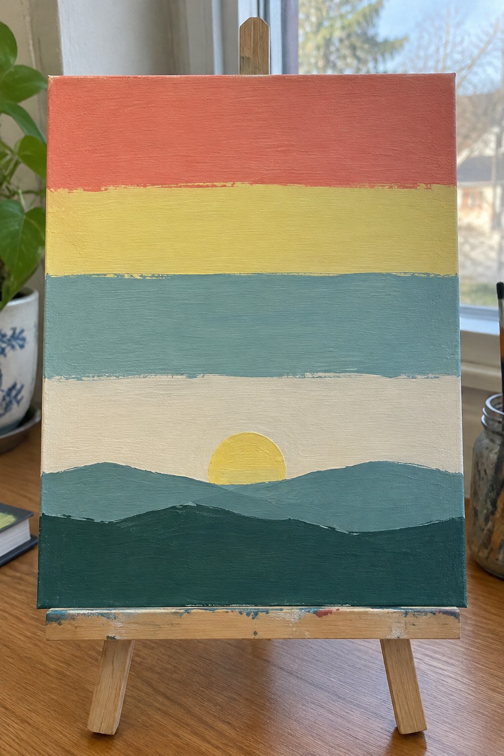 Layered sunset painting on easel with red yellow teal stripes and yellow sun over hills