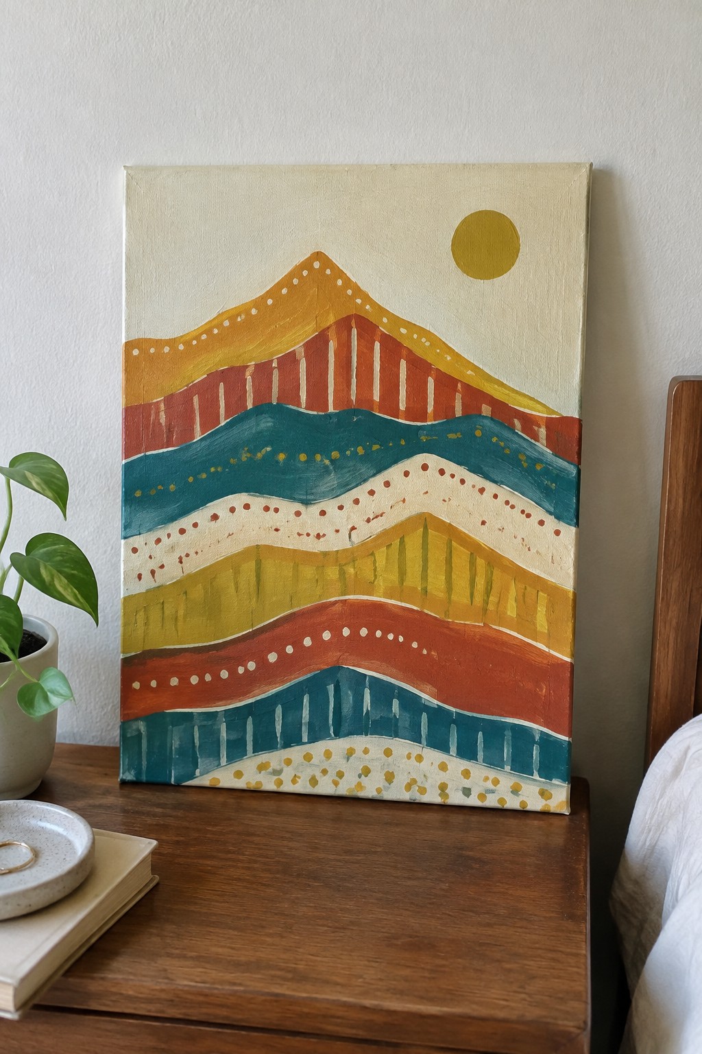 Colorful abstract painting of layered mountains and sun beside potted plant on wooden table