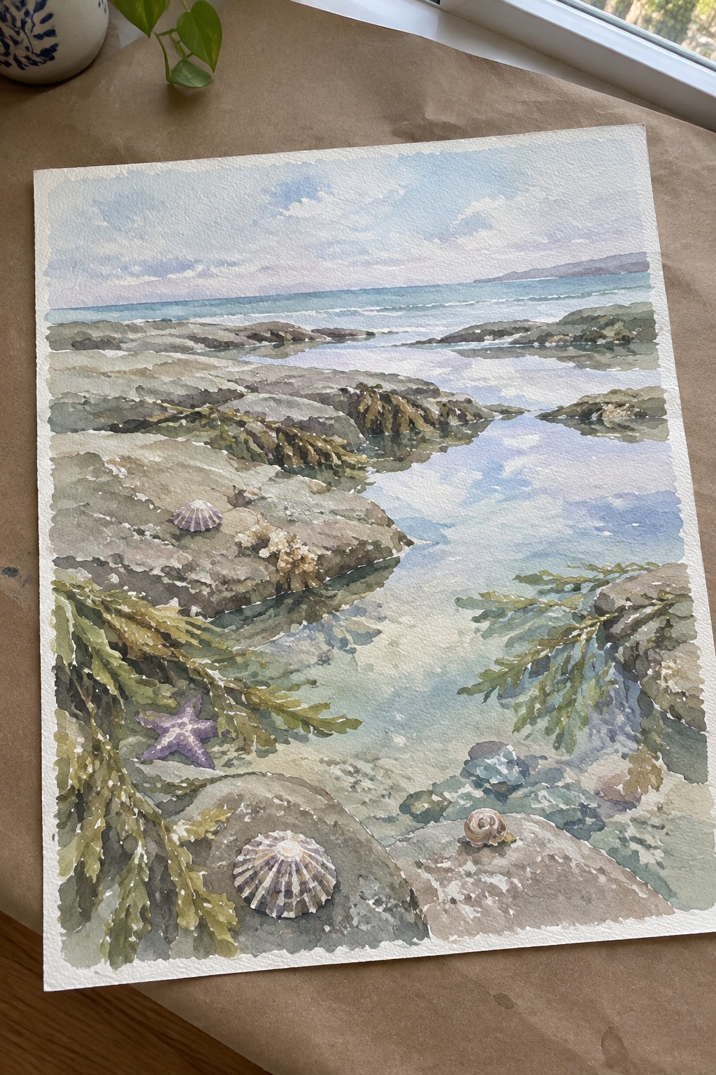 Watercolor seascape of rocky shore with purple starfish, seashells, seaweed, and ocean waves