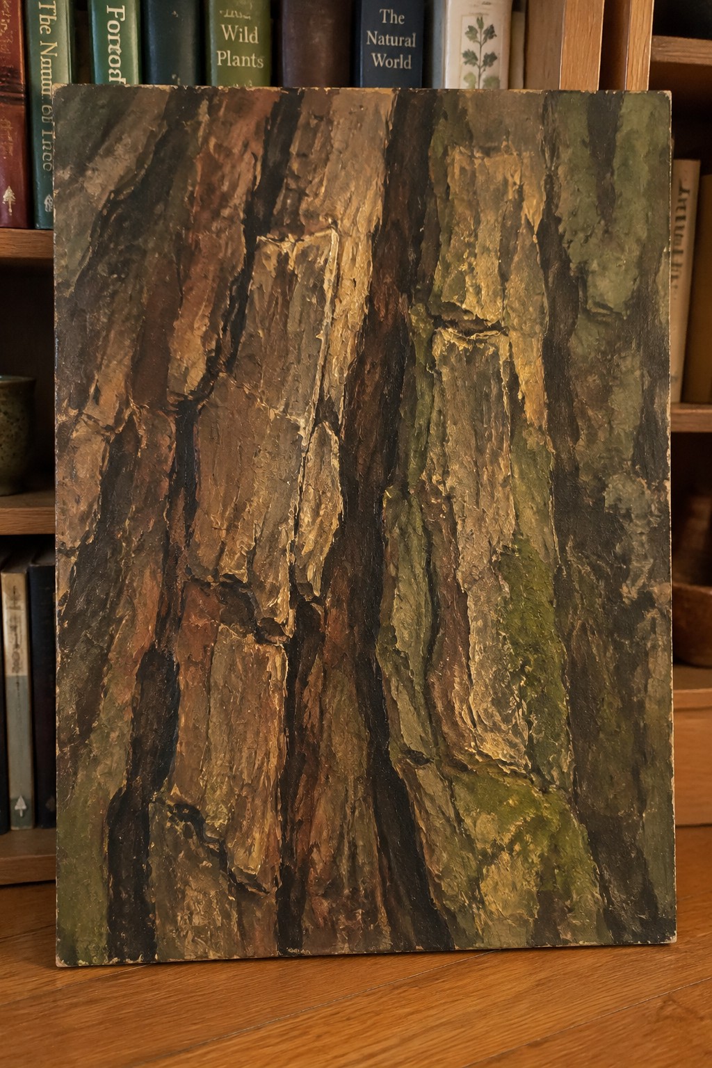 Close-up painting of rough tree bark with thick textured layers in brown, green, and gold tones leaning against a bookshelf.