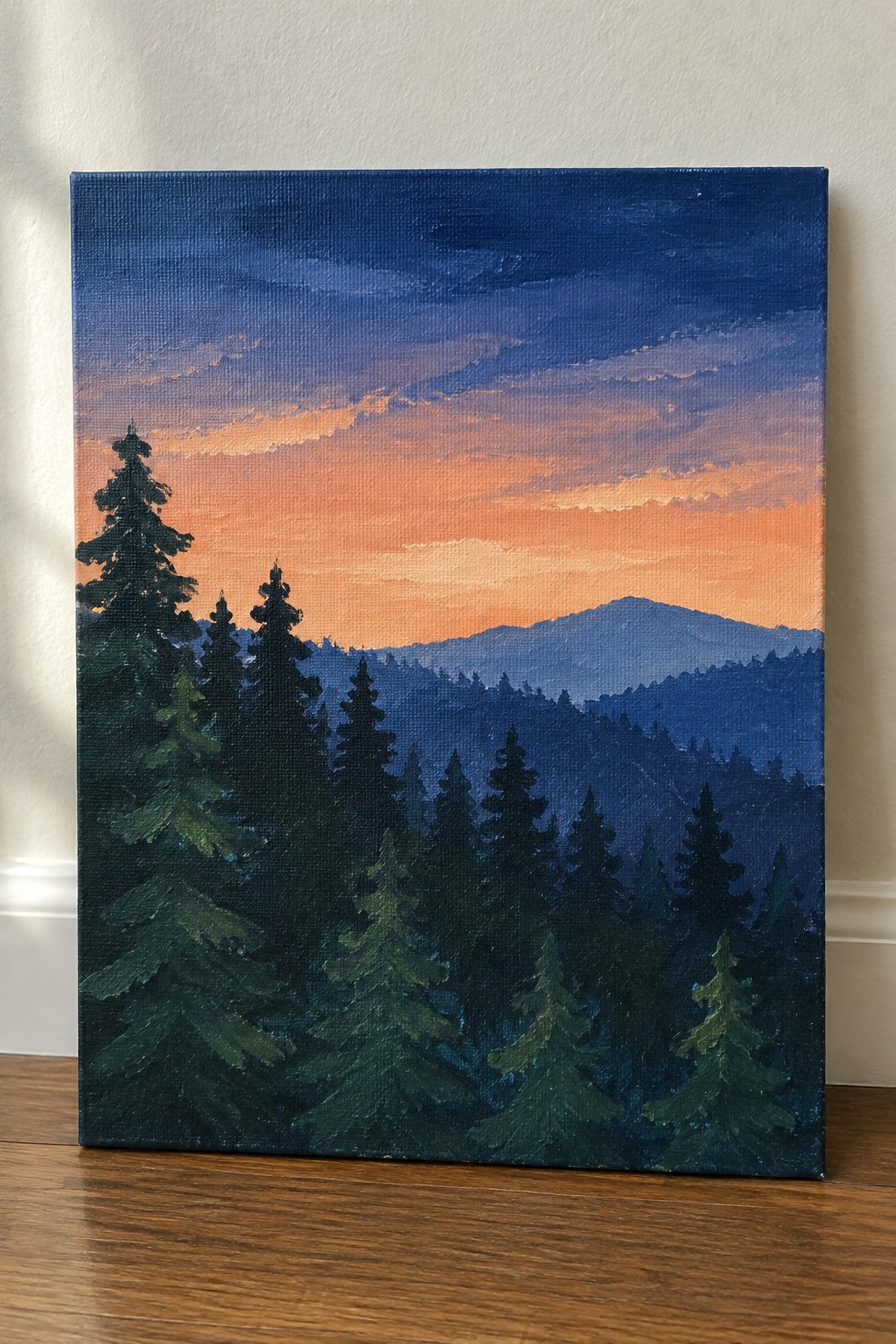 A rectangular canvas painting shows a dense pine forest silhouette in dark green and blue tones against a sunset sky that gradients from orange near the horizon to deep blue above, with layered mountains in the distance.