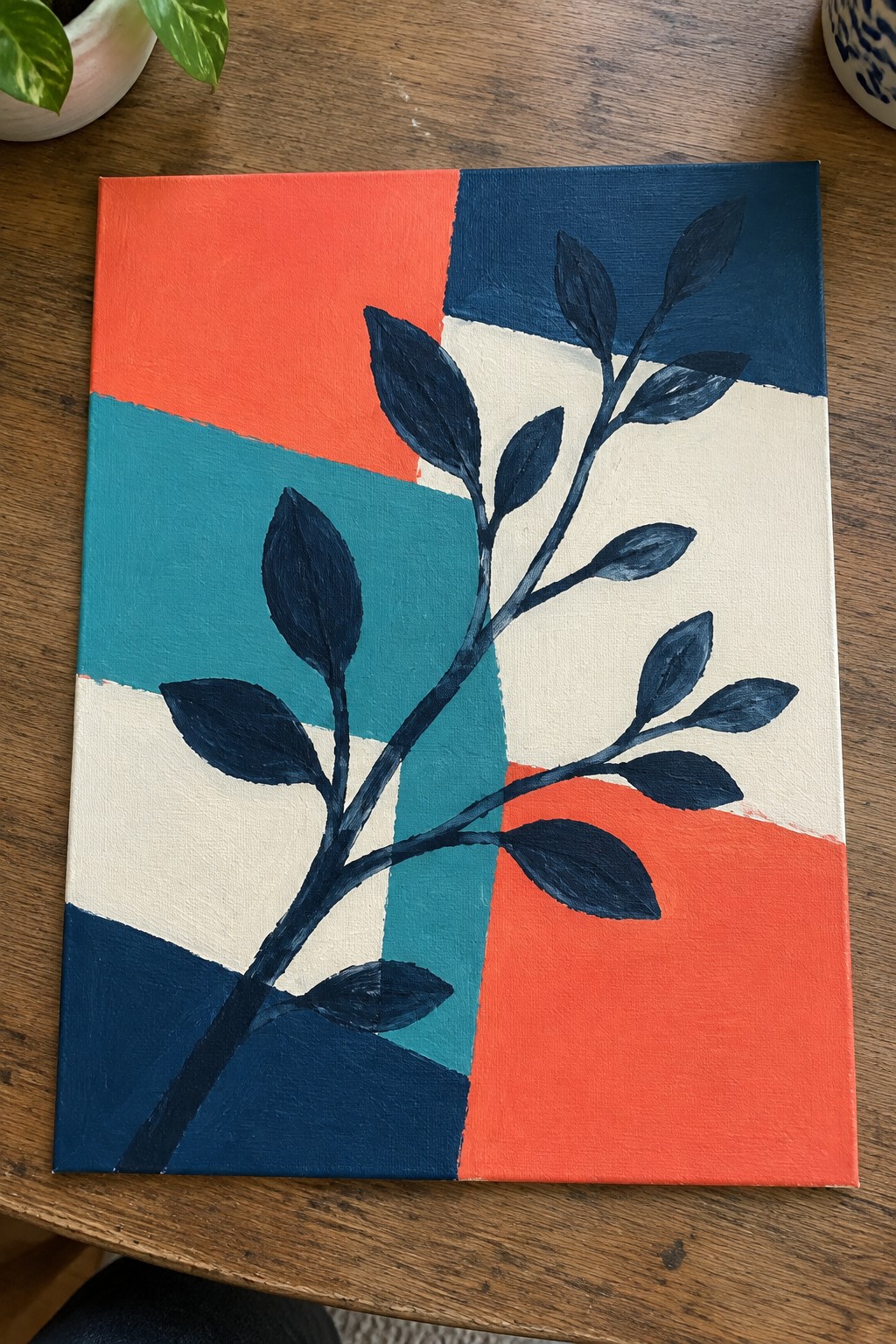 Abstract painting with coral, teal, navy blocks and dark leafy branch on wood table.