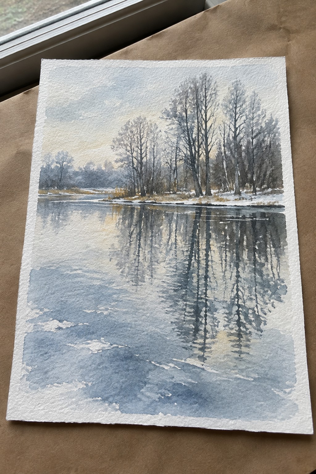 Watercolor of bare trees reflected in calm blue winter river with snowbanks.
