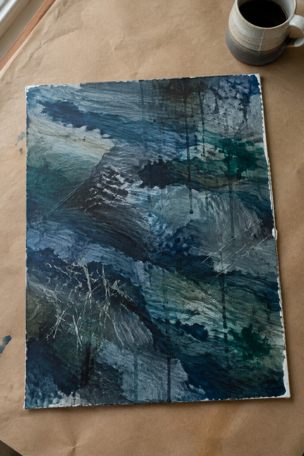 Abstract blue painting on canvas beside ceramic mug on brown paper.