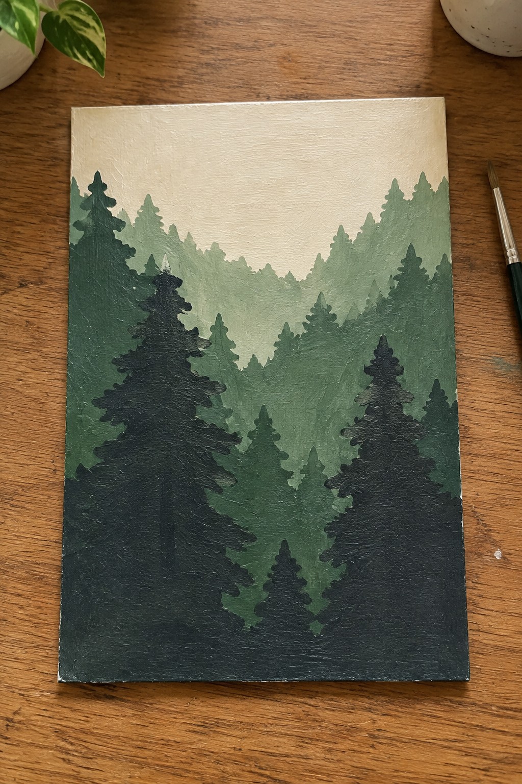 Layered green pine forest painting on canvas beside paintbrush on wood table