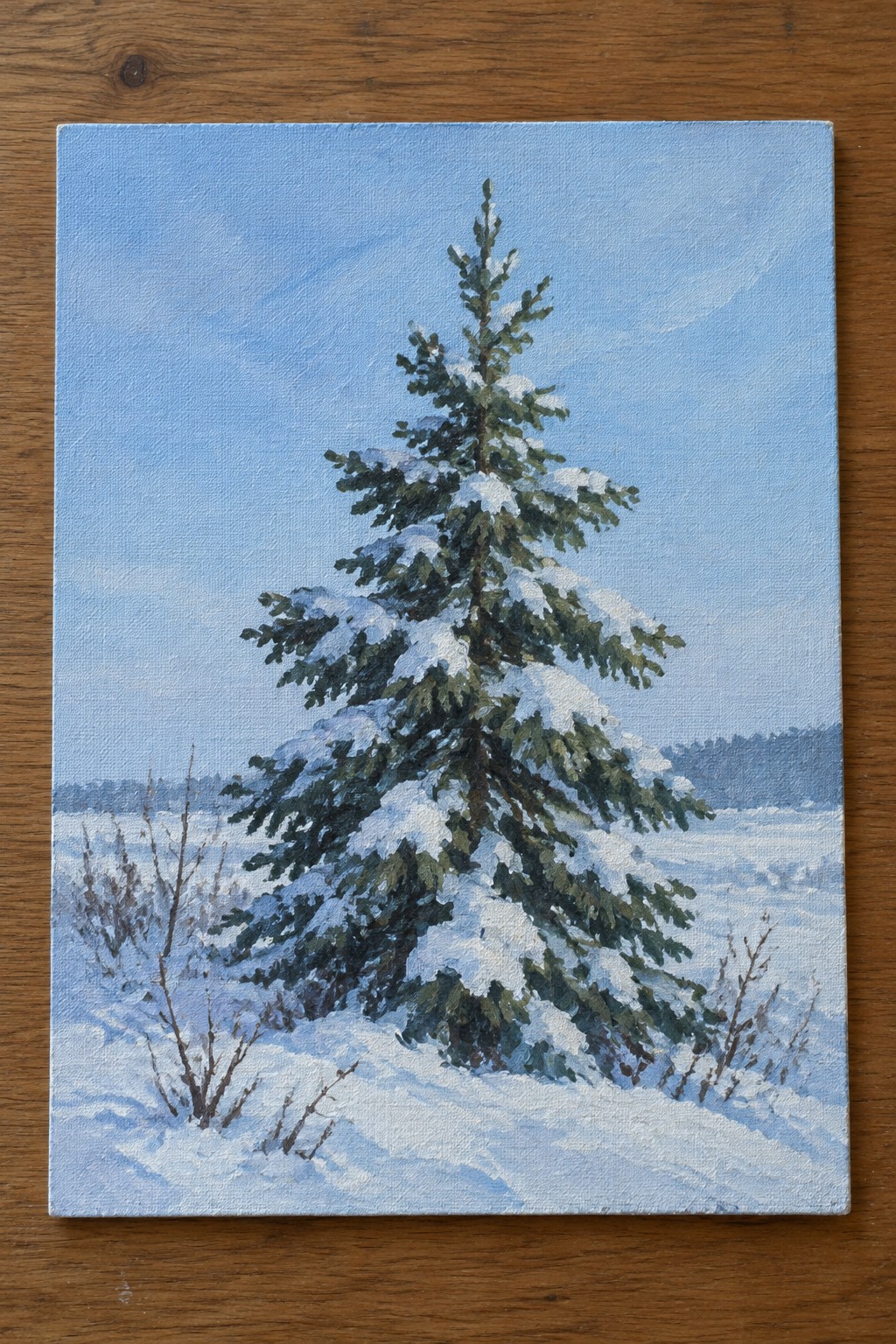 Snow-covered pine tree in oil painting on canvas atop wooden table