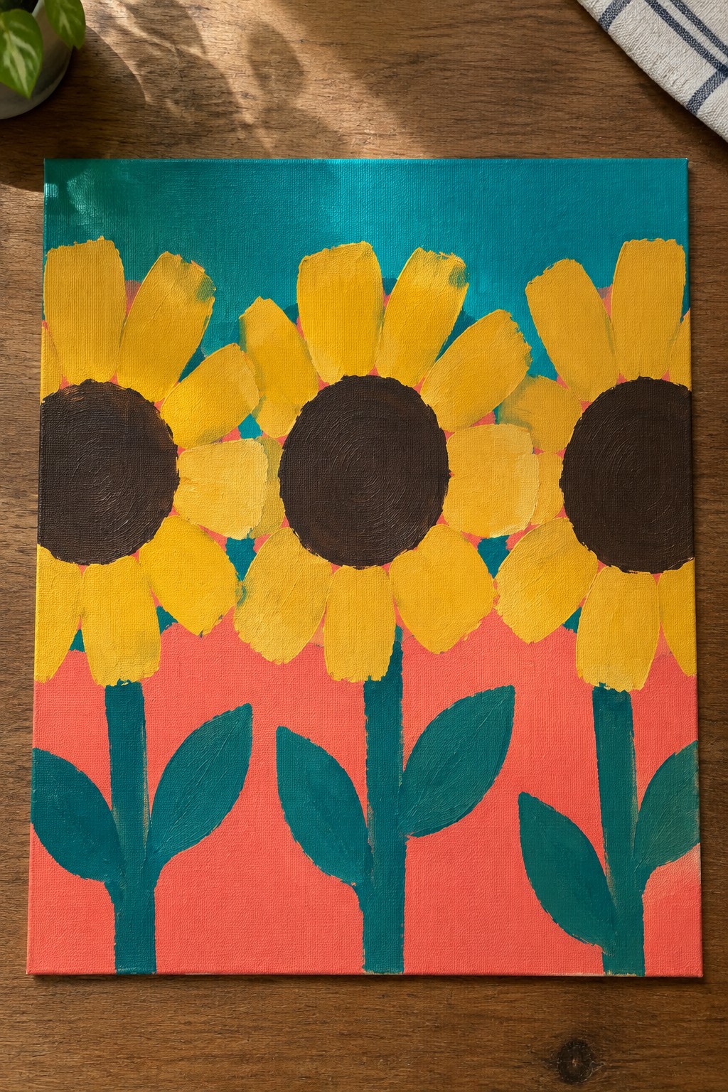 A square canvas painting shows three sunflowers with yellow petals and dark centers above green stems and leaves, set against a teal upper background and coral lower background.