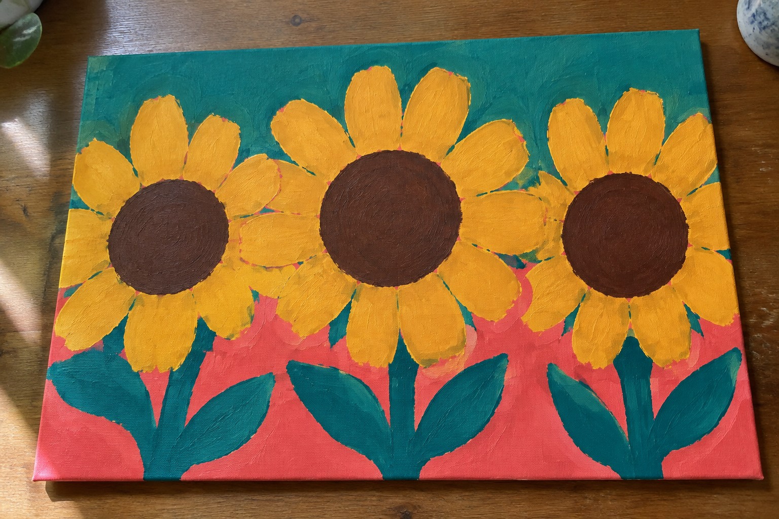 A square canvas painting shows three sunflowers with yellow petals and dark centers above green stems and leaves, set against a teal upper background and coral lower background.