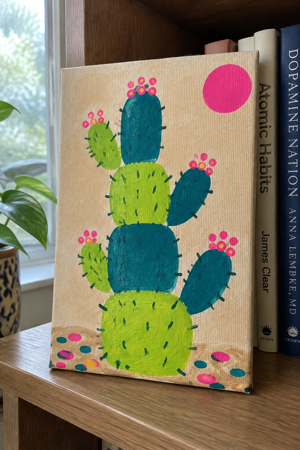 Colorful cactus painting with pink flowers and sun on canvas beside books on shelf