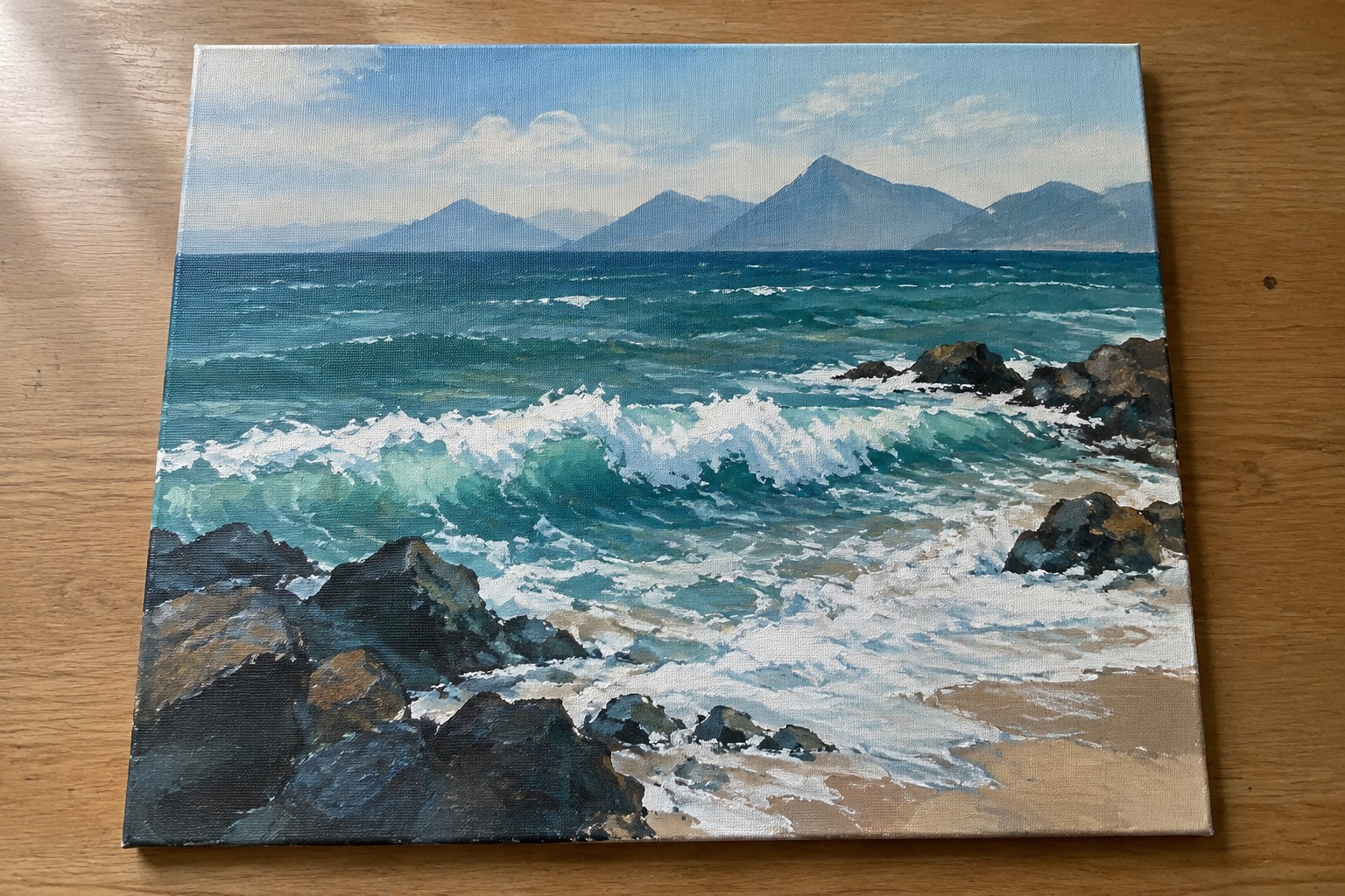 An acrylic painting of turquoise waves with white foam crashing on dark rocks along a sandy beach, with layered mountains under a blue sky in the background.