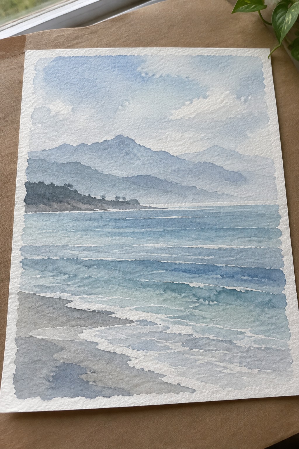 Watercolor seascape of layered blue mountains above wavy ocean meeting a rocky shore.