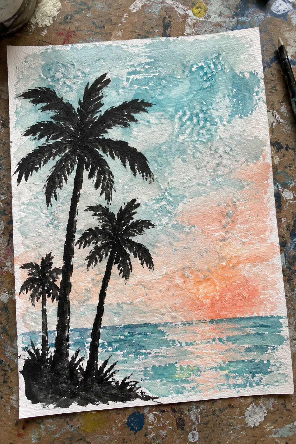 Watercolor painting of three black palm tree silhouettes by a turquoise ocean at sunset