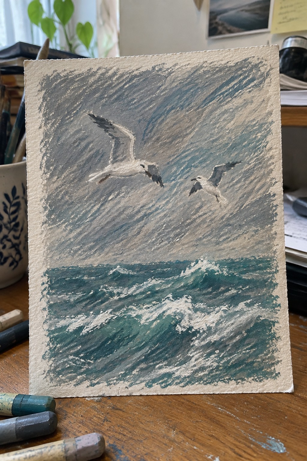 Pastel drawing of two seagulls over churning teal waves beneath a stormy gray sky