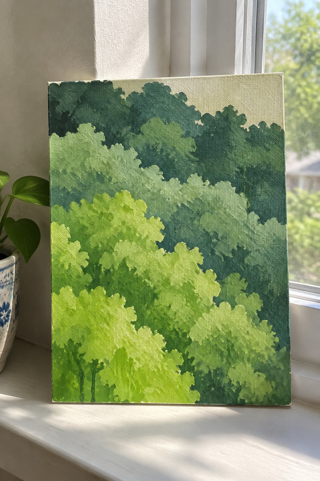 Green forest foliage painting on sunlit windowsill beside potted plant with heart-shaped leaves