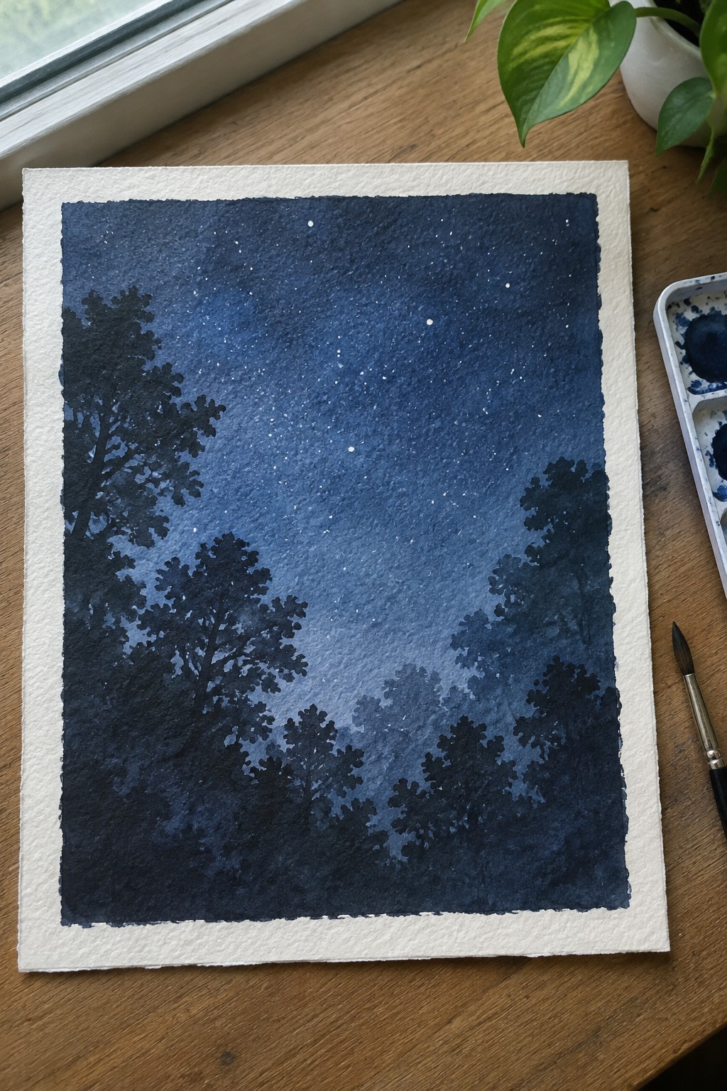 A dark blue watercolor night sky with white stars and black tree canopy silhouettes painted on textured paper next to a palette and brush.