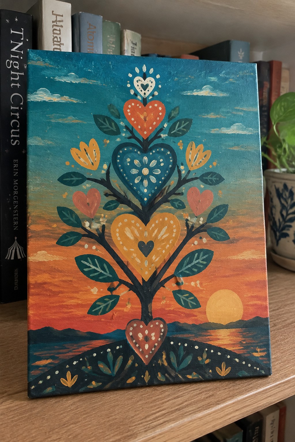 Folk art painting of heart-shaped tree with colorful leaves against vibrant sunset over water