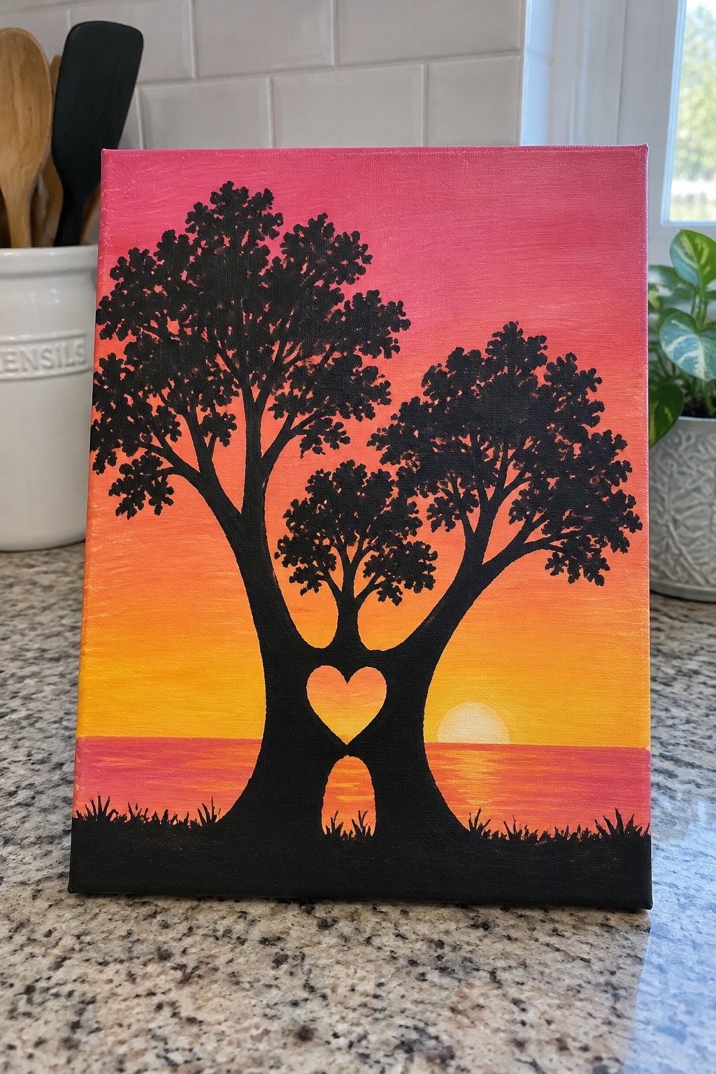 A canvas shows two black tree silhouettes forming a heart shape where the trunks meet, painted against a pink-to-orange sunset gradient with a small sun on the horizon and grass at the bottom.