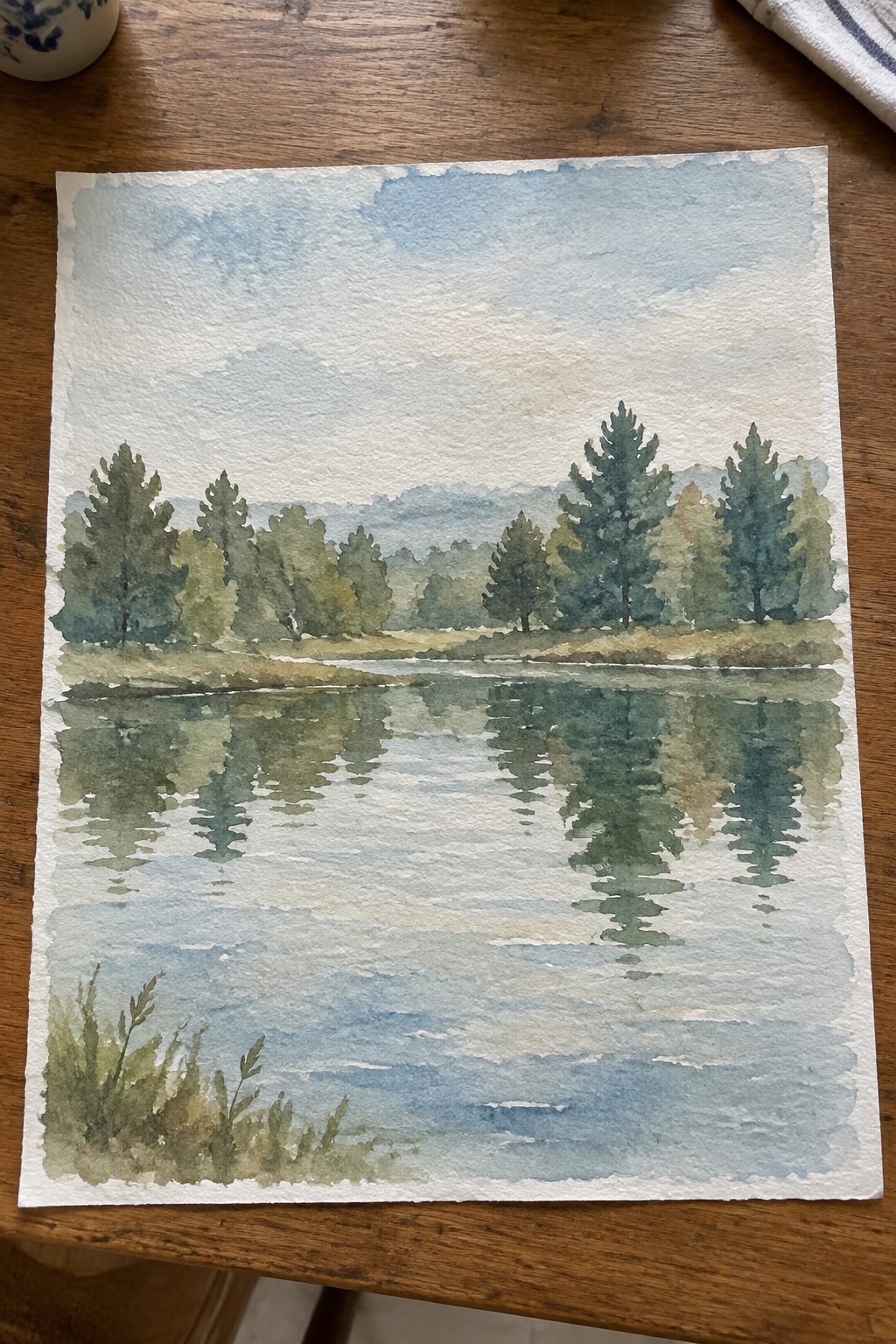Watercolor landscape of reflective lake with pine trees and hills on wooden table