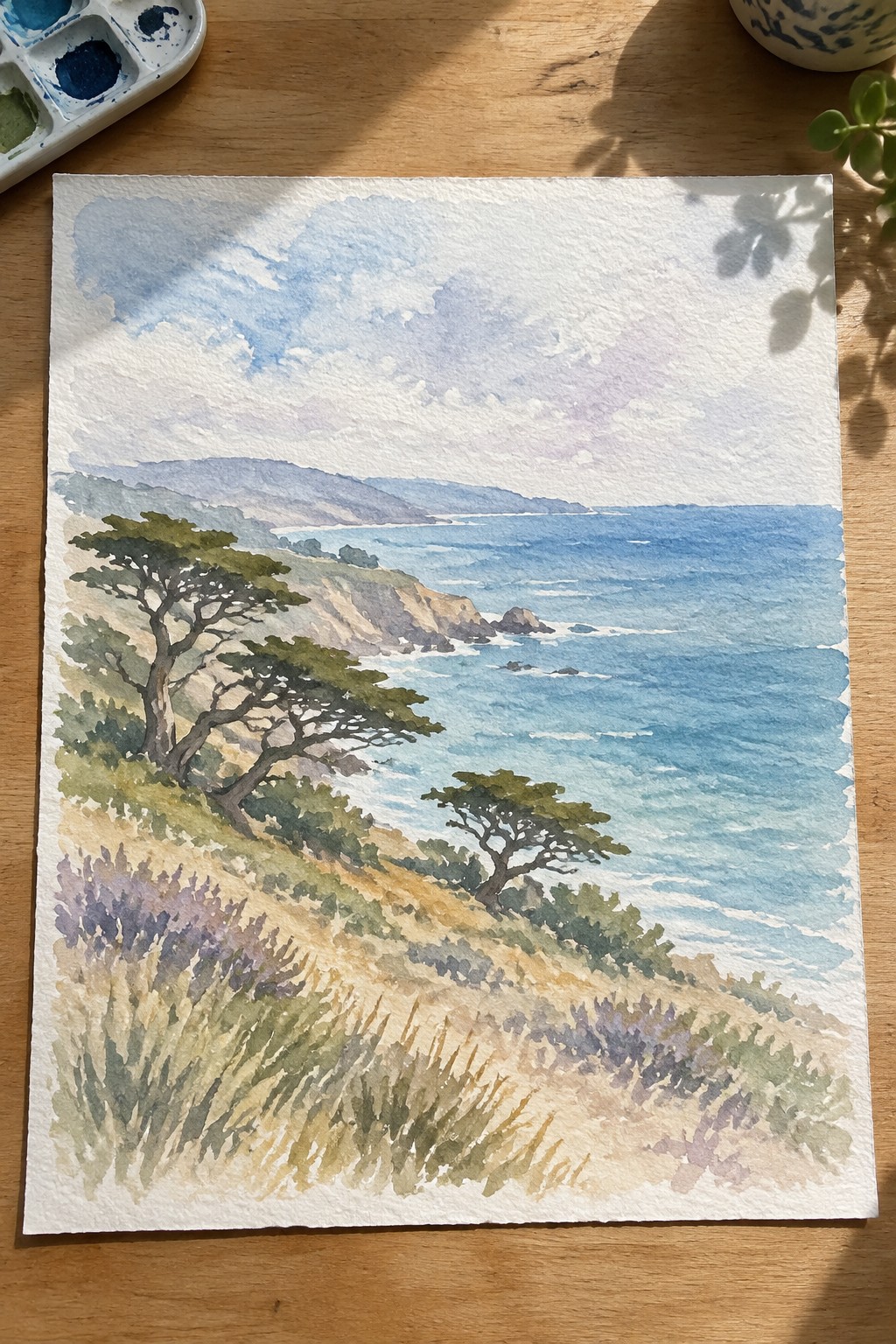 Watercolor coastal scene with cliffs, trees, blue ocean, palette on wooden table