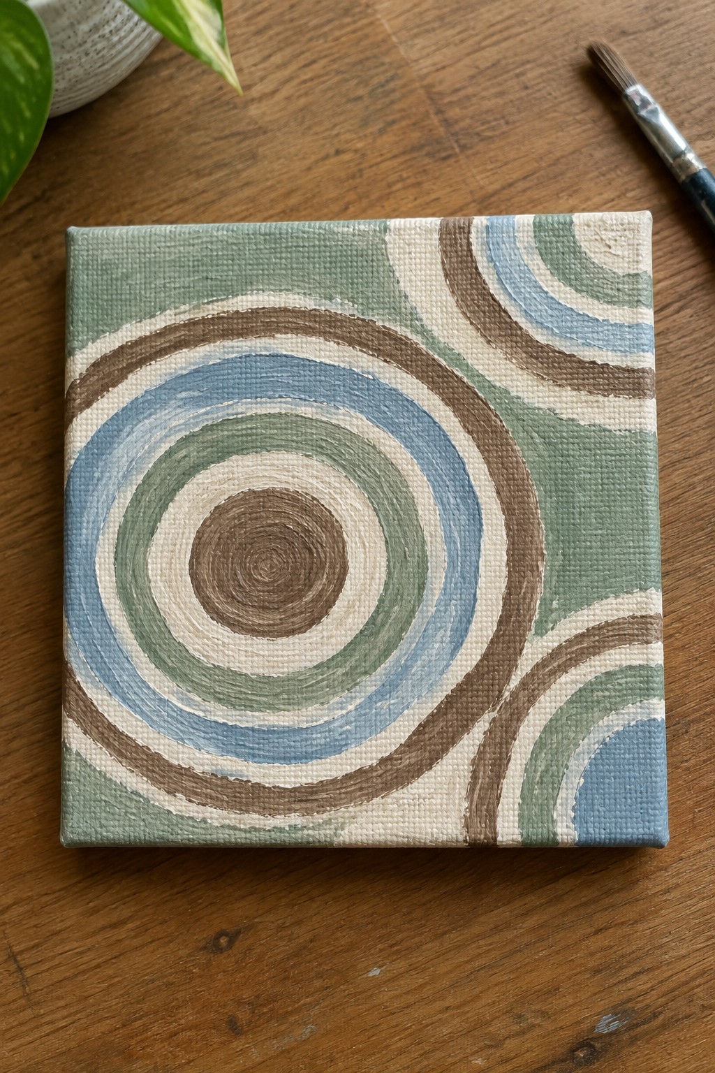 A small square canvas with overlapping concentric circles in blue, green, brown, and cream on a light green background sits on a wooden table next to a paintbrush.