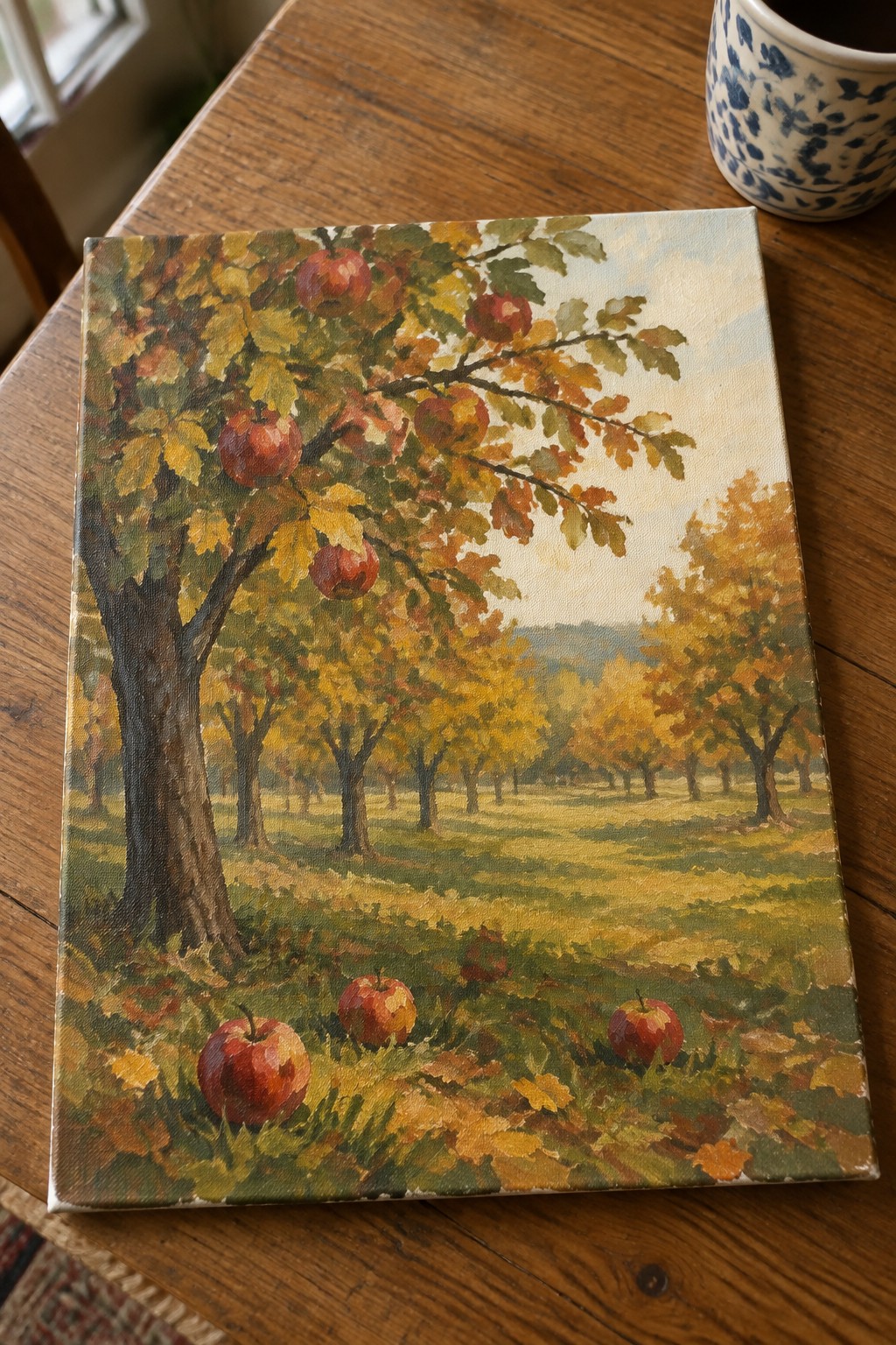 Autumn apple orchard painting on wooden table beside blue floral mug