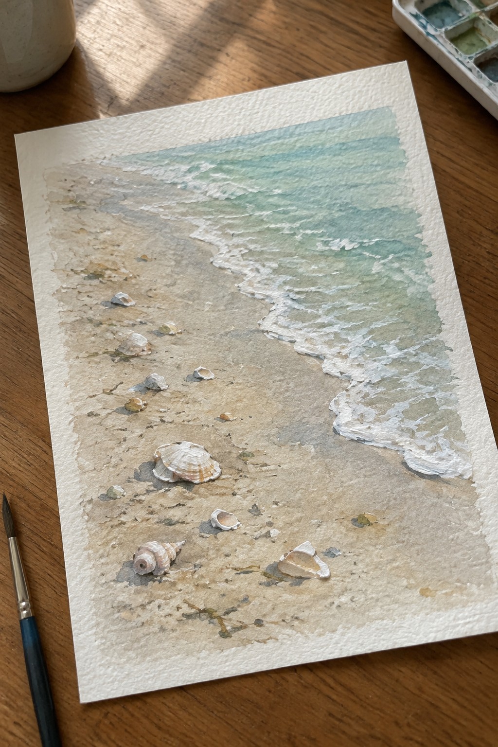 Watercolor painting of beach with seashells, turquoise waves, brush, and palette on wood.