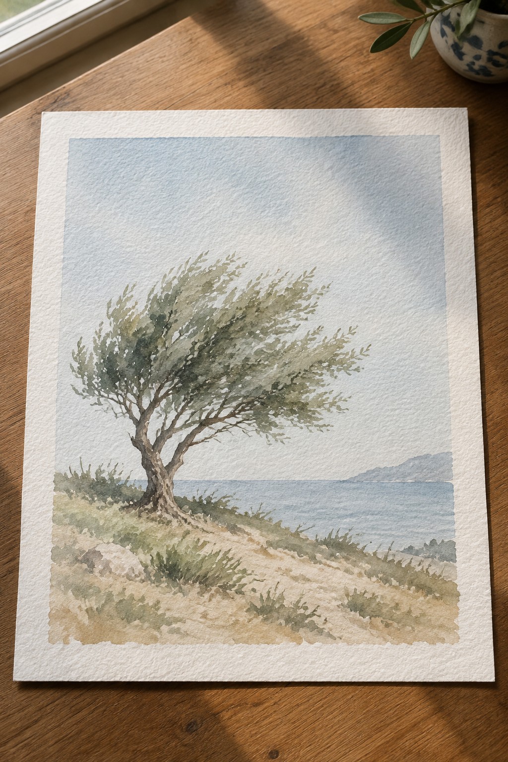 Watercolor painting of windswept tree overlooking blue sea on wooden table