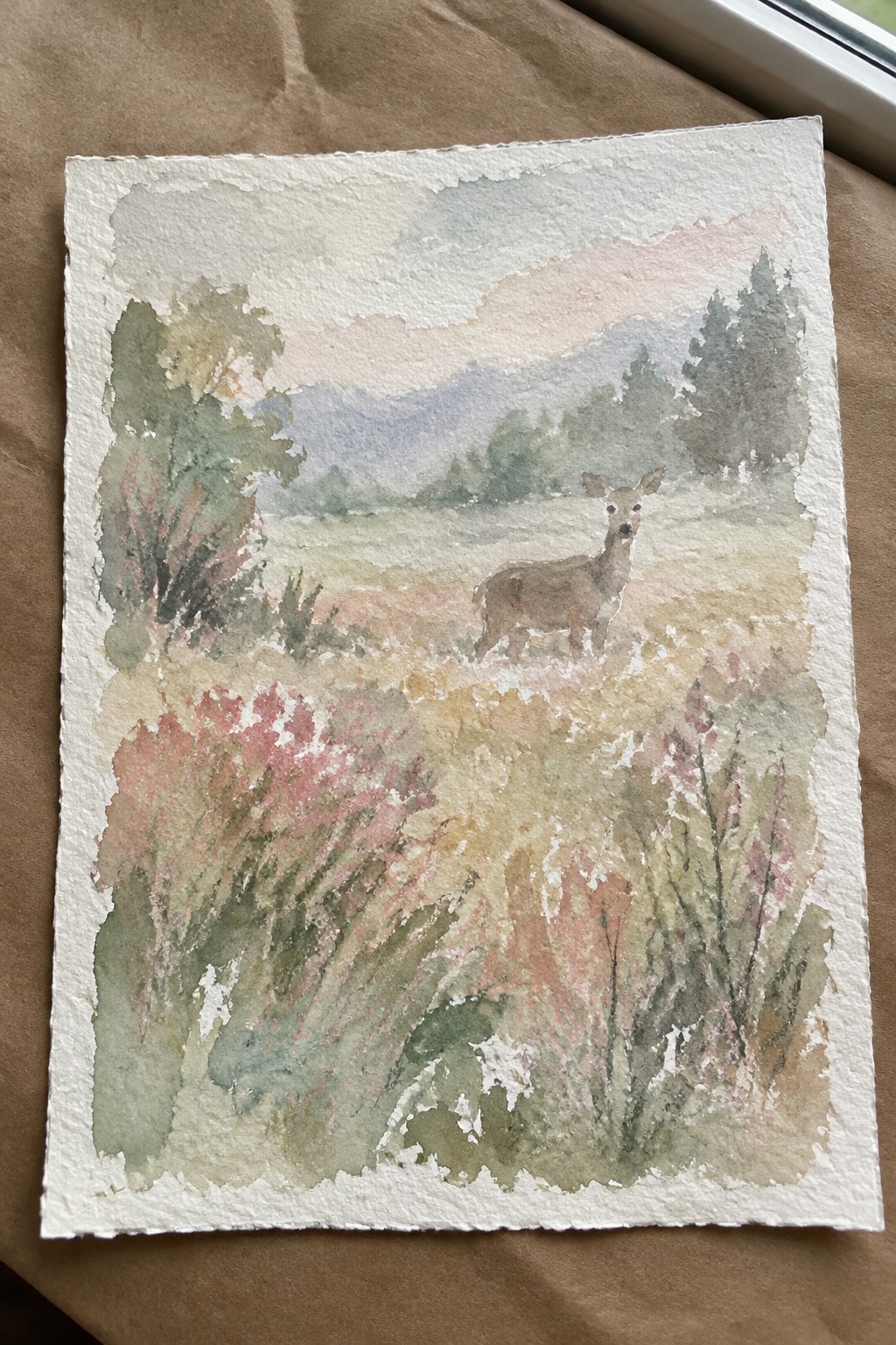 Watercolor painting of deer amid pink wildflowers in meadow with hazy mountains beyond