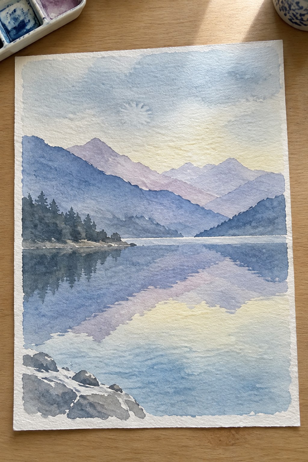 Watercolor painting of blue mountains reflected in a calm lake under a yellow sky.
