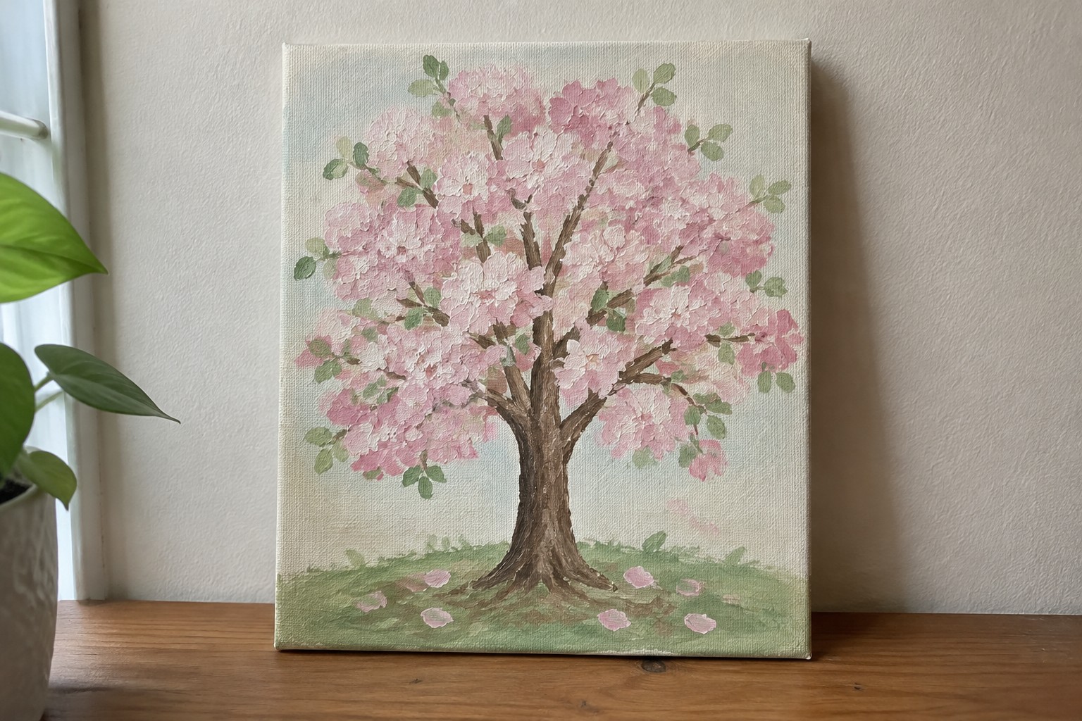 A canvas painting of a cherry blossom tree with pink flowers, brown trunk, and green leaves sits on a wooden surface beside a potted plant.