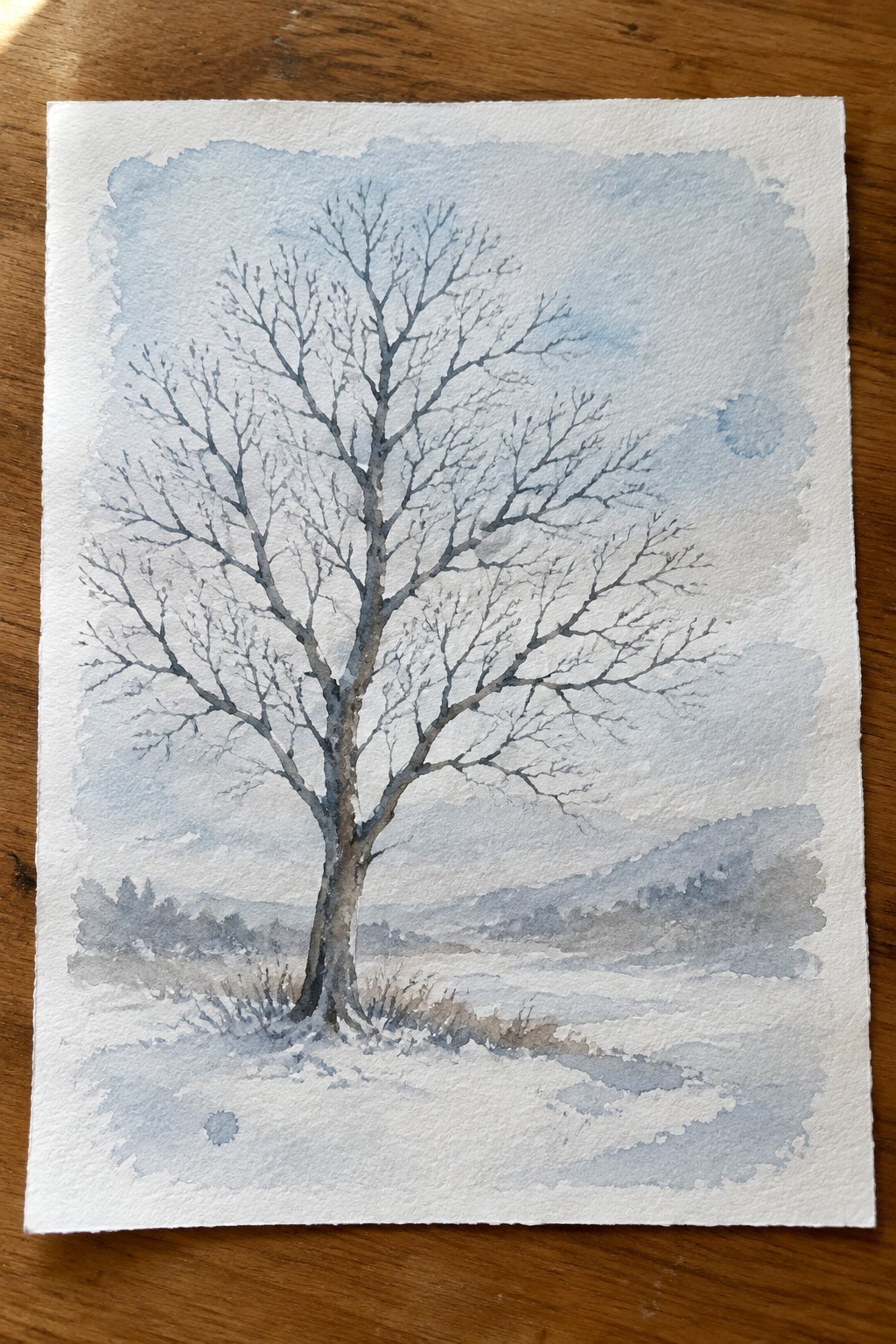 Watercolor of bare tree in snowy field with misty hills under pale blue sky