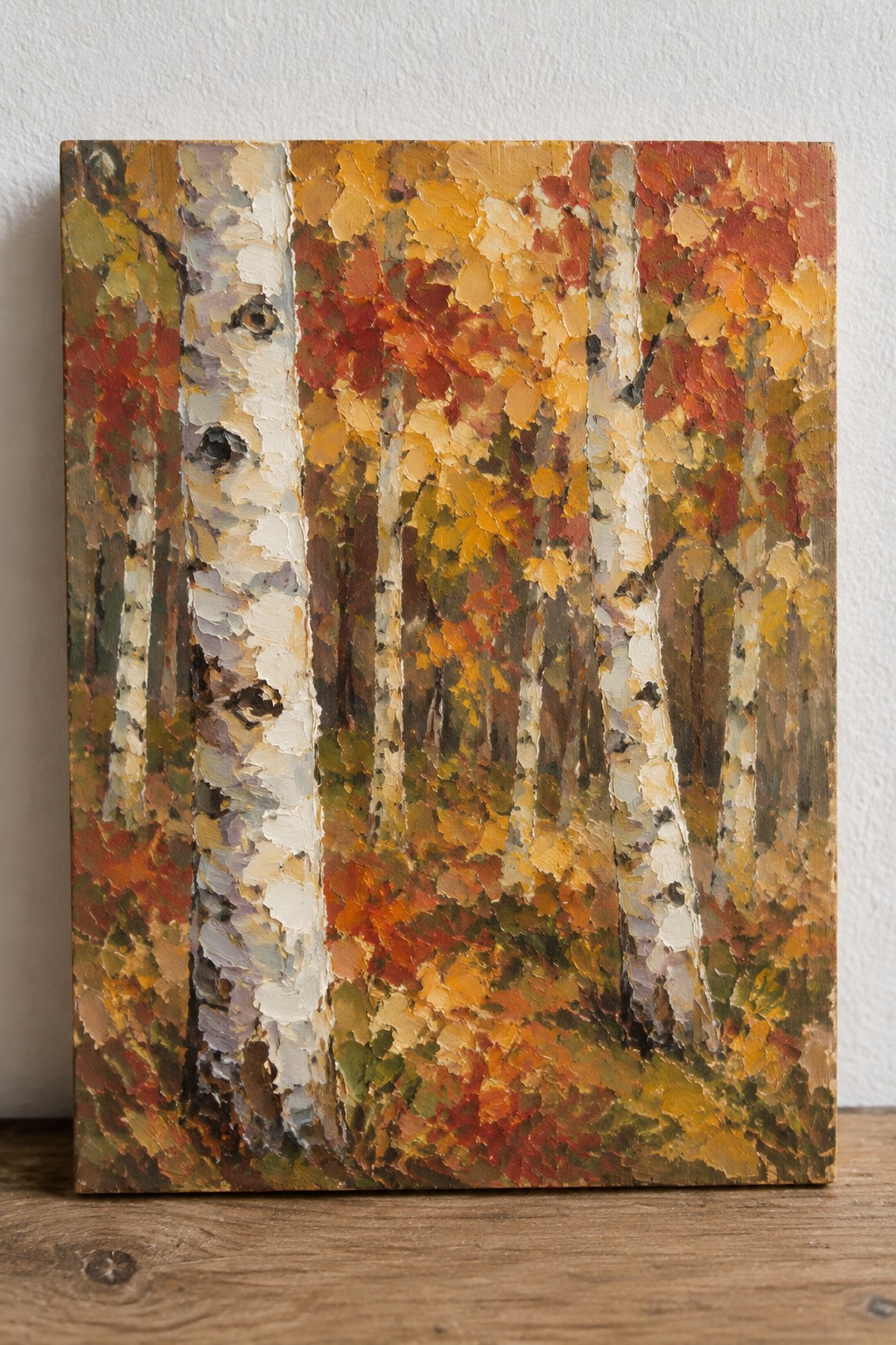 An oil painting of several birch trees with white trunks and black markings, surrounded by thick layers of red, orange, and yellow autumn leaves on a wooden surface.