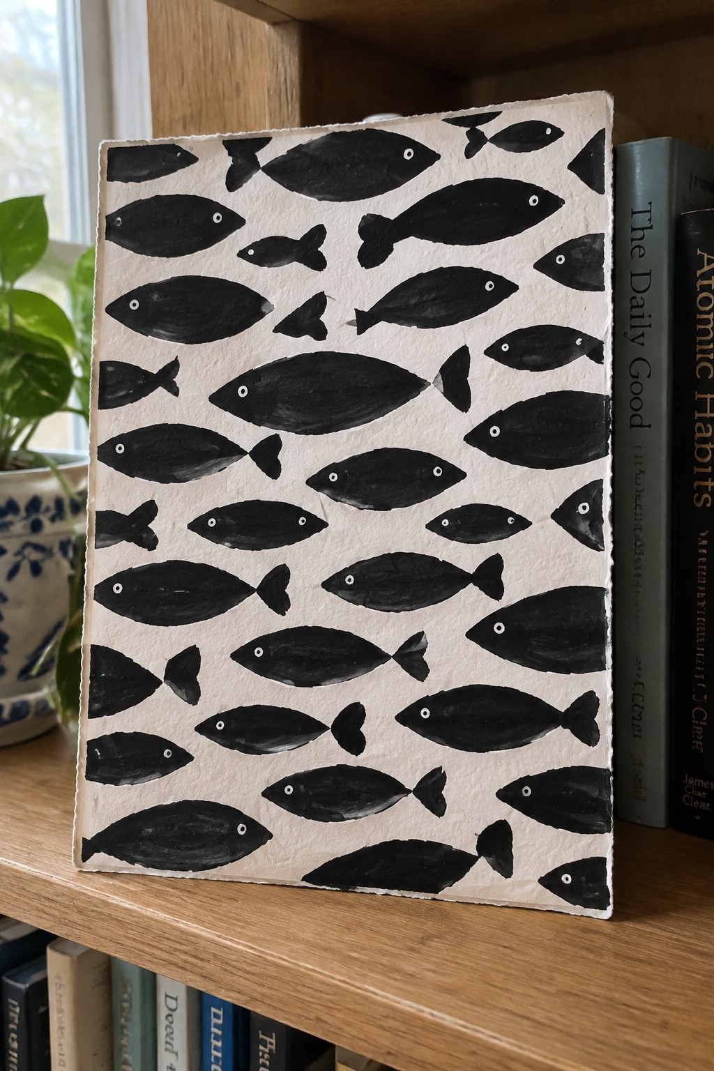 Hand-painted black fish on white textured paper on wooden bookshelf with books