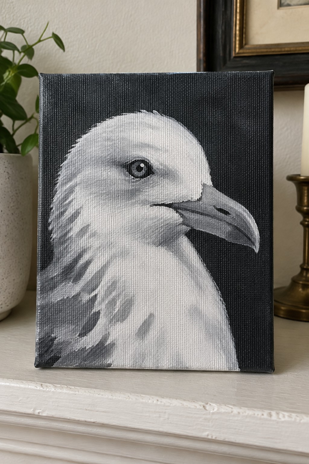 Black canvas painting of white seagull head on white mantel with plant and candle