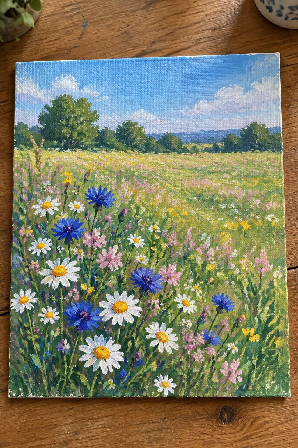 Vibrant canvas painting of wildflower meadow with white daisies, blue cornflowers, and green trees