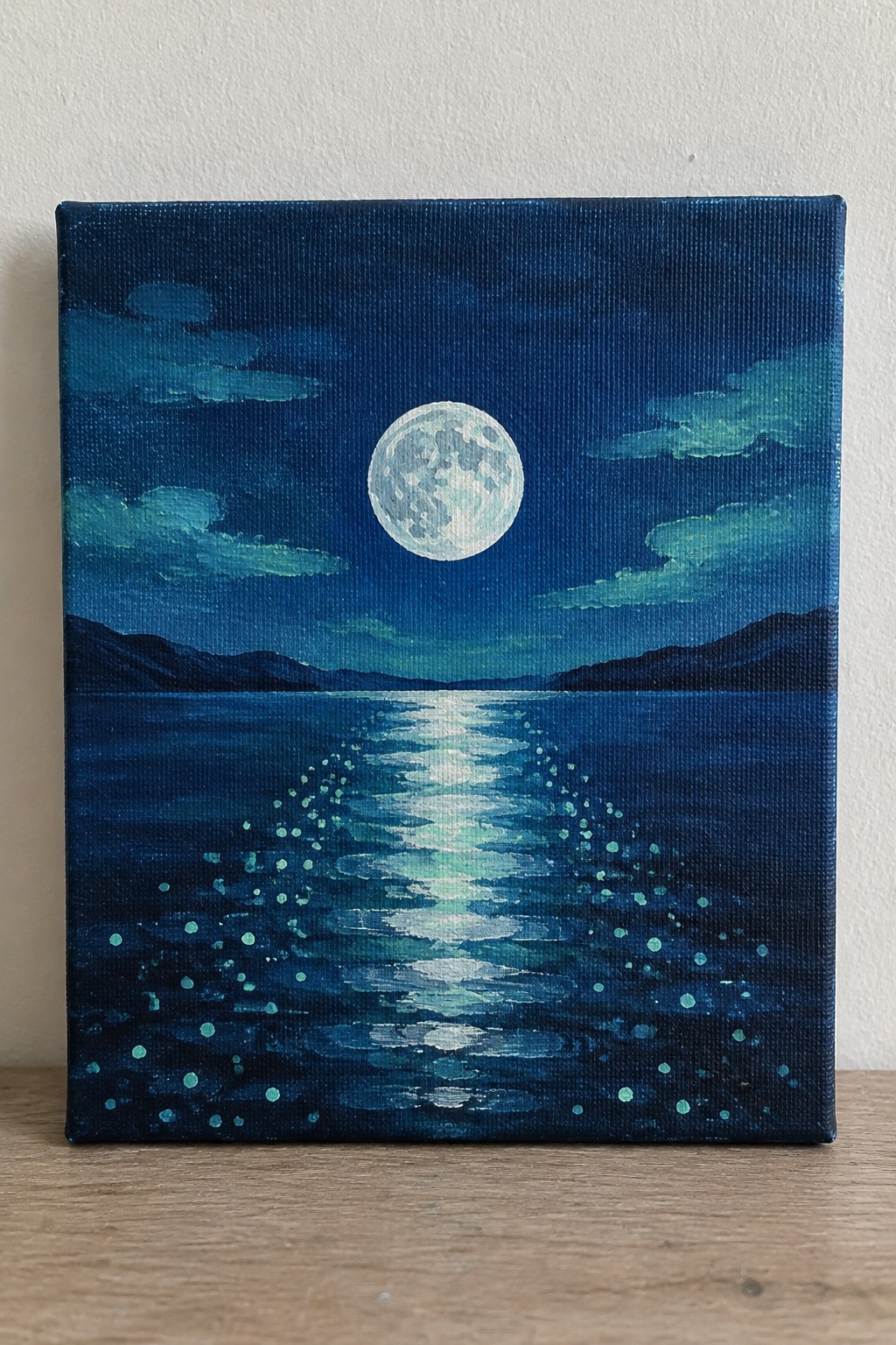 Full moon shines above dark blue waters with glowing reflection path and silhouetted mountains.