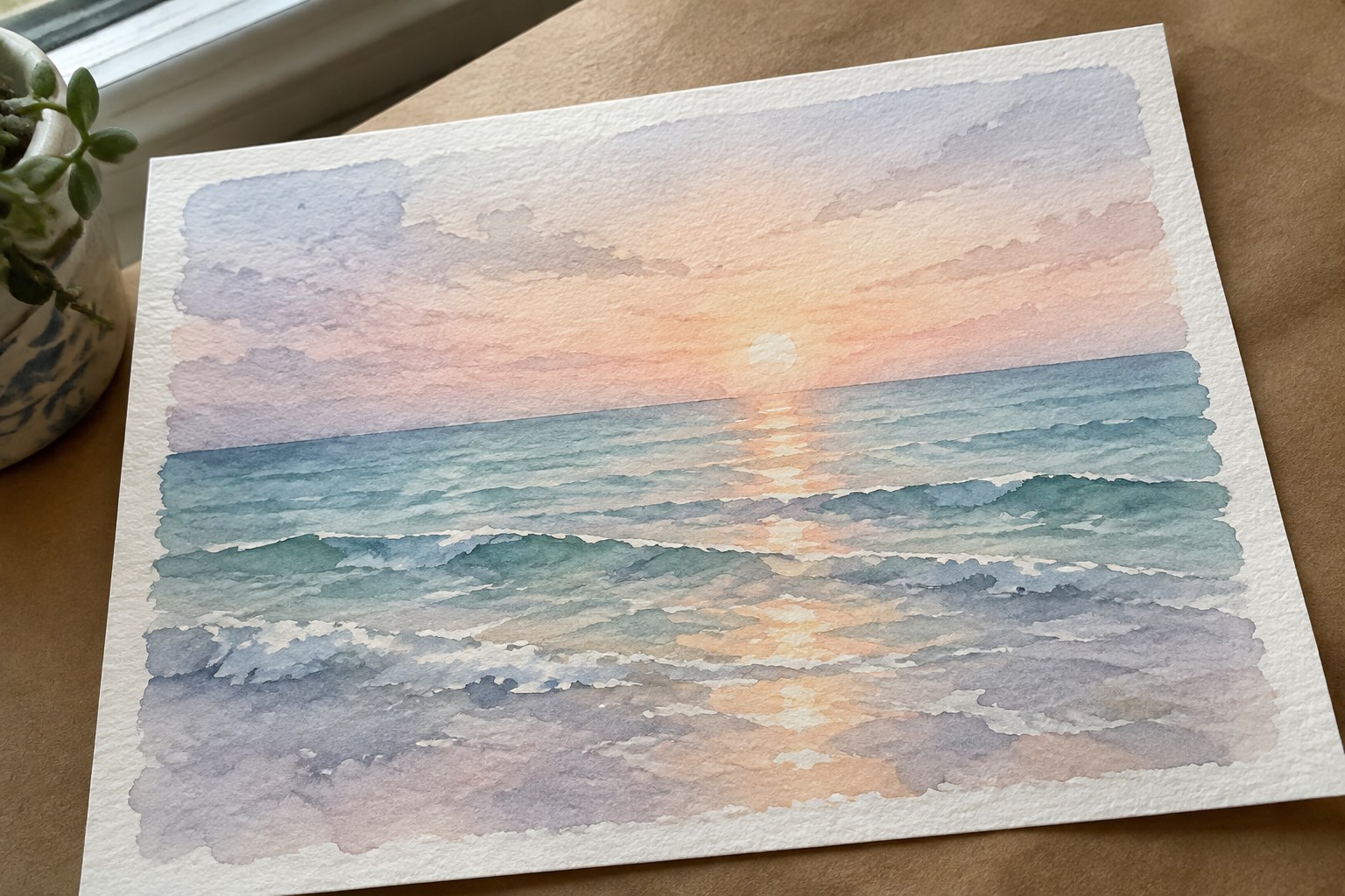 A watercolor painting of a sunset over the ocean with waves and a sun reflection on the water, placed on a brown surface next to a plant.