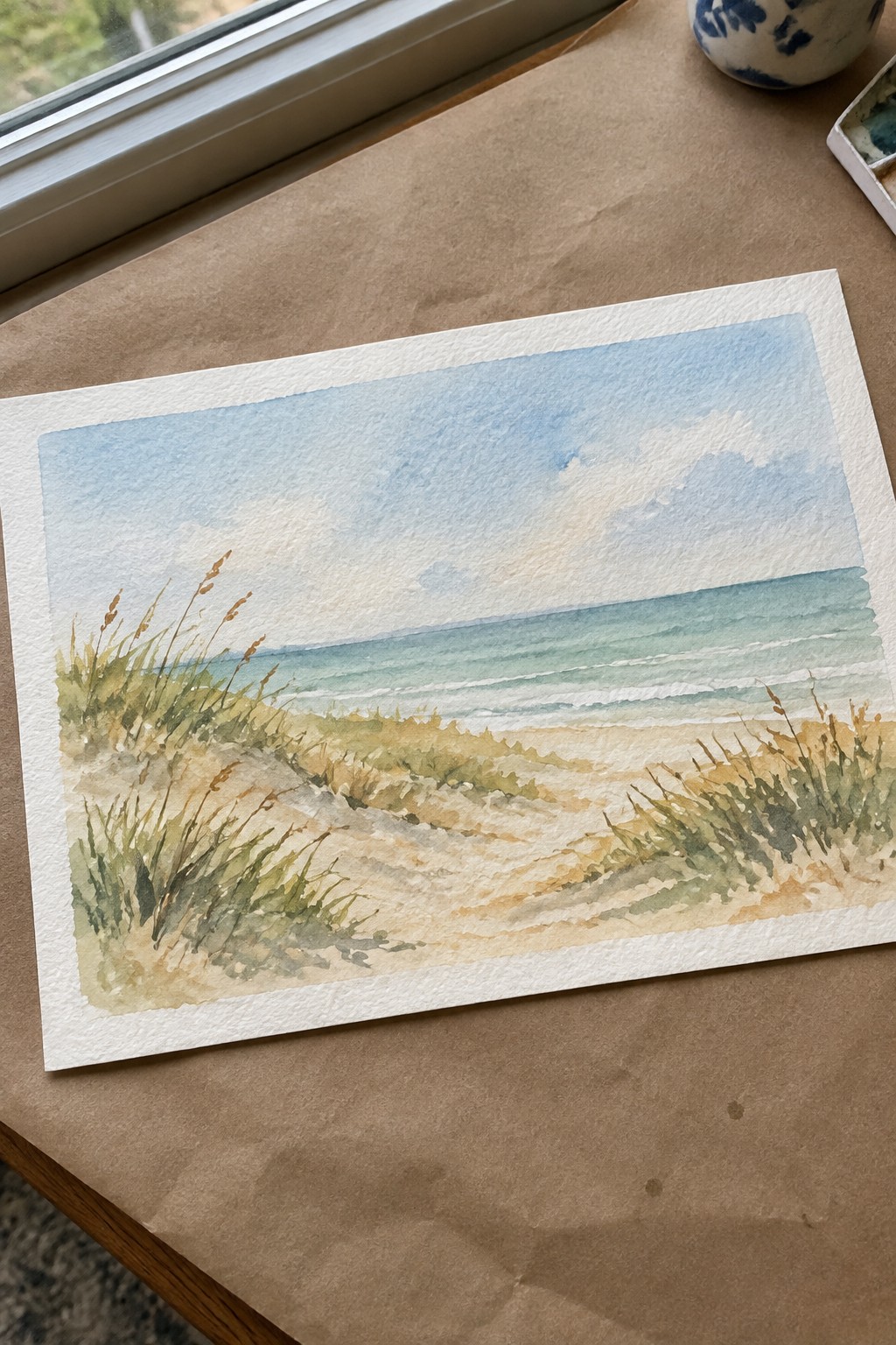 A watercolor painting of a beach scene with sandy dunes and tall grass in the foreground leading to a calm ocean under a pale blue sky.