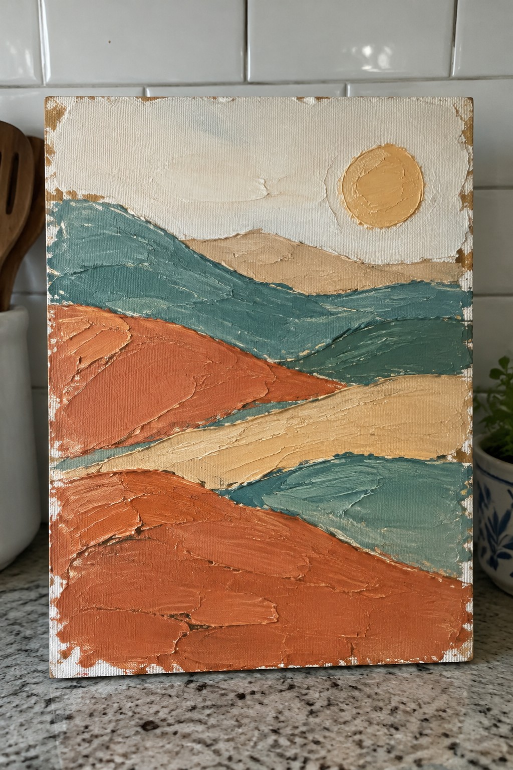 Abstract painting of layered orange, teal, and beige hills with yellow sun on kitchen counter.